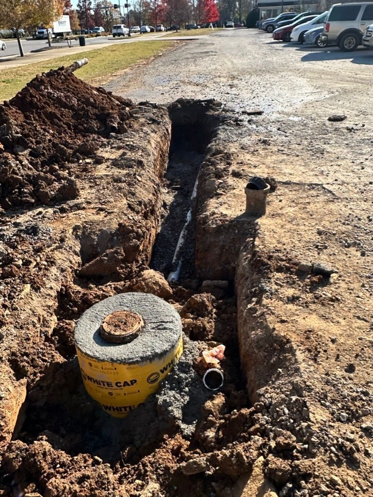 Open trench beside a parking lot, with dirt piles and a yellow utility cap in the foreground.