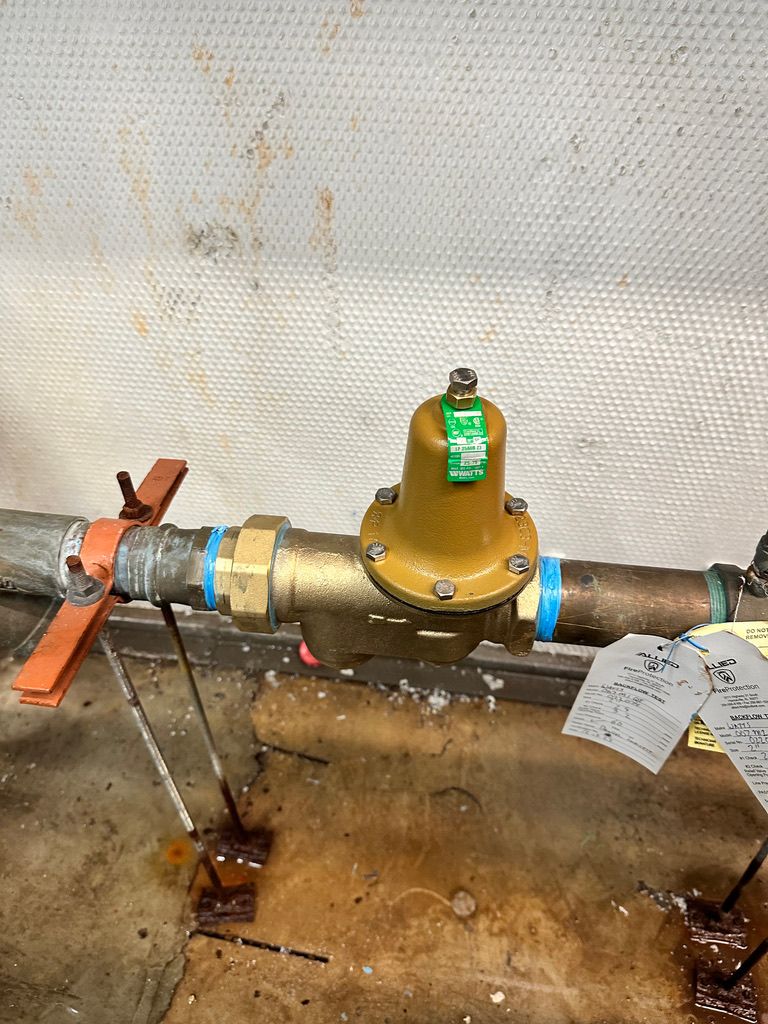 Brass pipe valve assembly with green cap on a wall-mounted pipe in a utility area, with water stains below