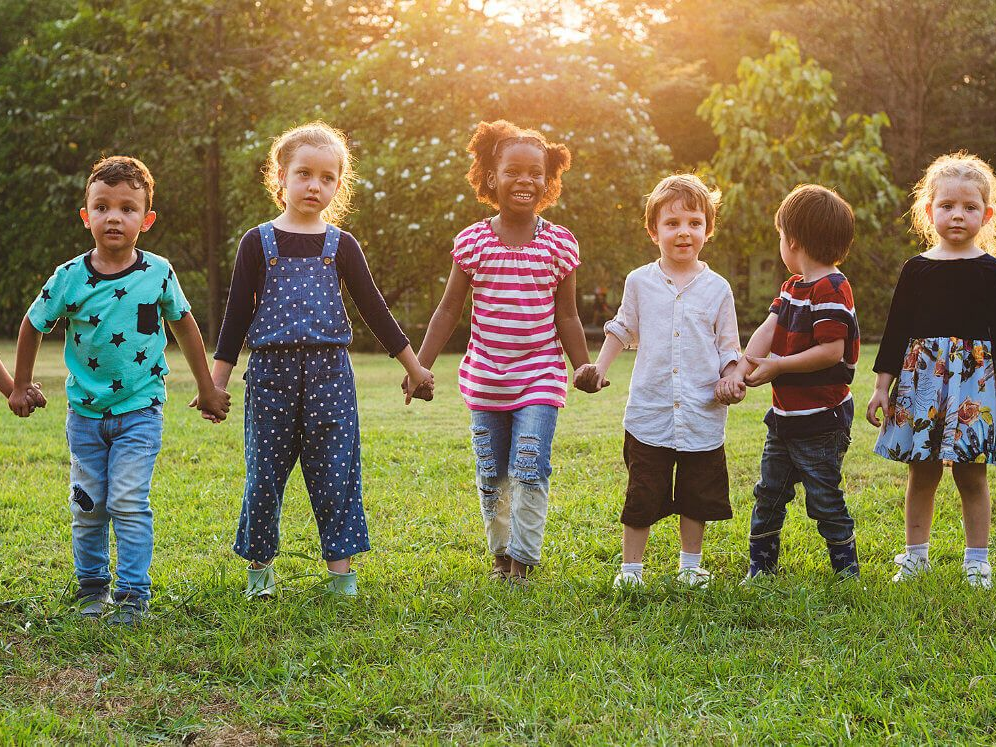 Seven children holding hands, standing on grass in a sunny park.