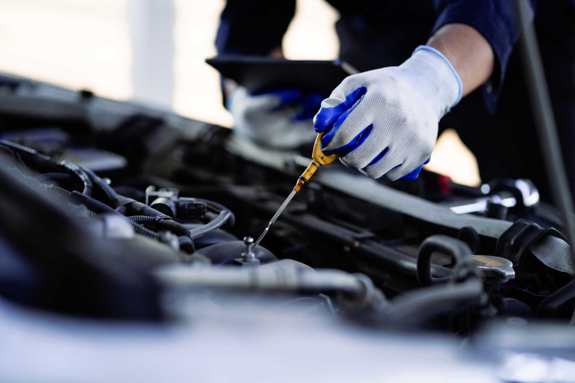 Mechanic checking car oil with dipstick, wearing gloves, holding tablet.