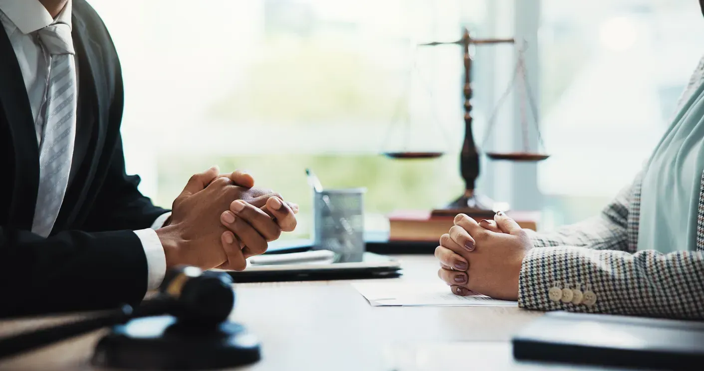 Two people in business attire discuss across a table, with a scales of justice blurred in the background.