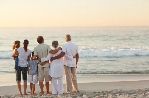 Five people standing arm-in-arm on a beach at sunset, facing the ocean.