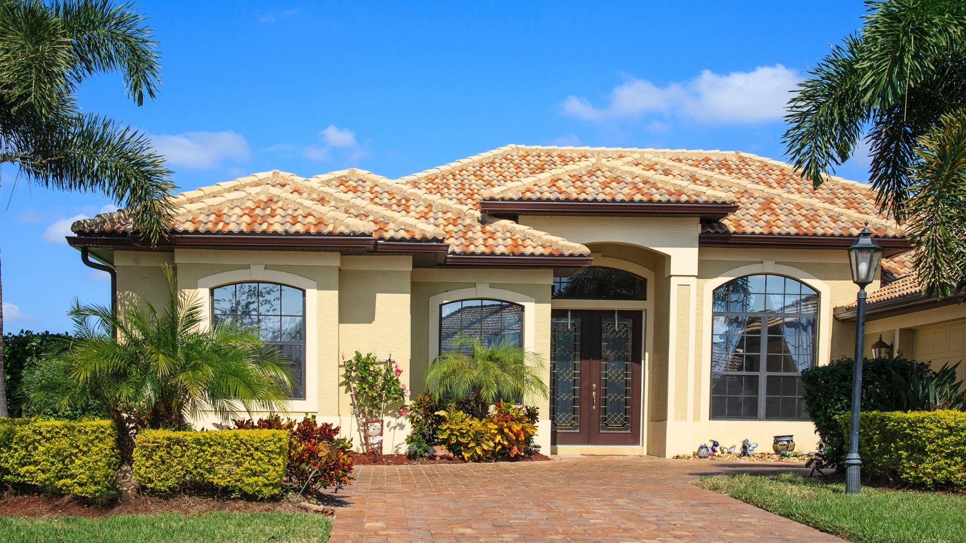 Single-story Mediterranean-style house with tan stucco, terracotta roof, arched windows, and a brick driveway.