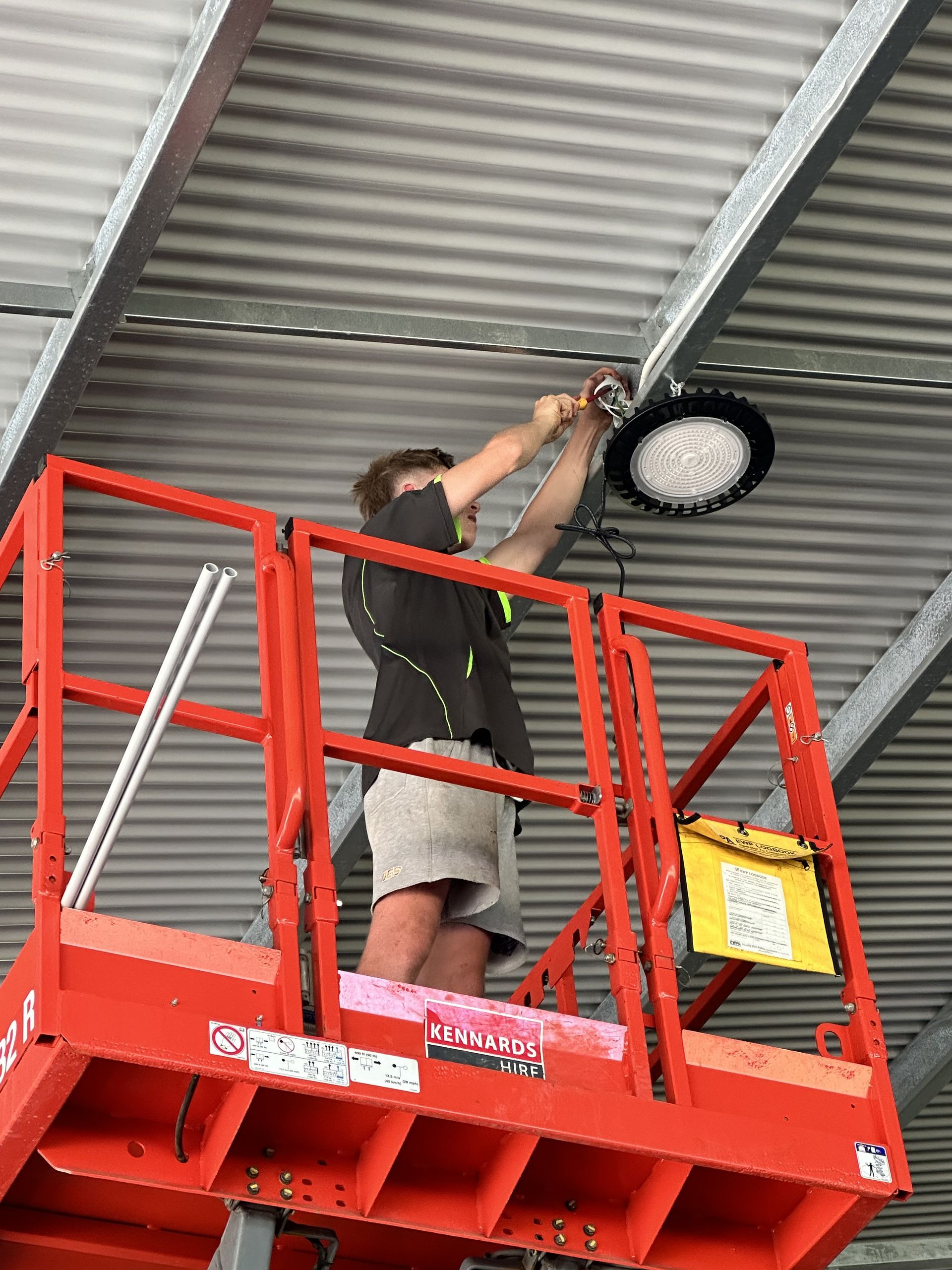 Person on a red lift installing a large circular light fixture in a warehouse. — DG Electrical & Fire In Kiama, NSW