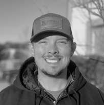 A man wearing a hat and a hoodie is smiling in a black and white photo.
