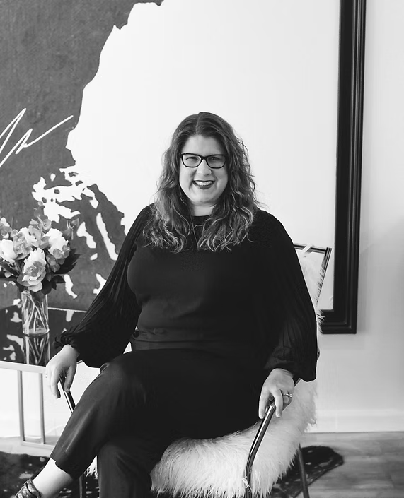 Woman in glasses sits in a chair, smiling, against a black and white art piece, wearing all black.