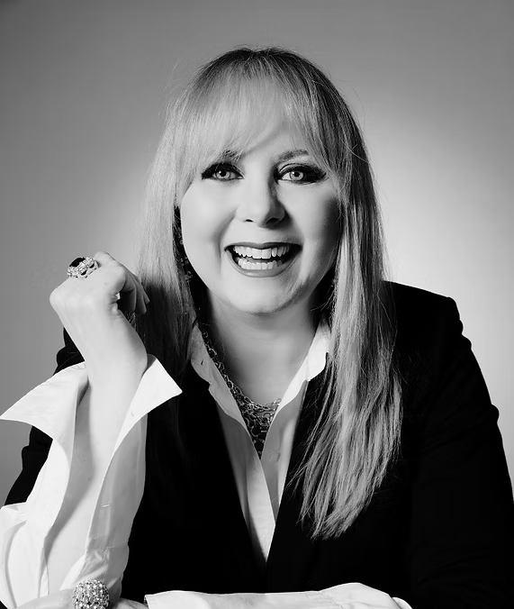 Woman in black blazer smiles, resting arm on chin.