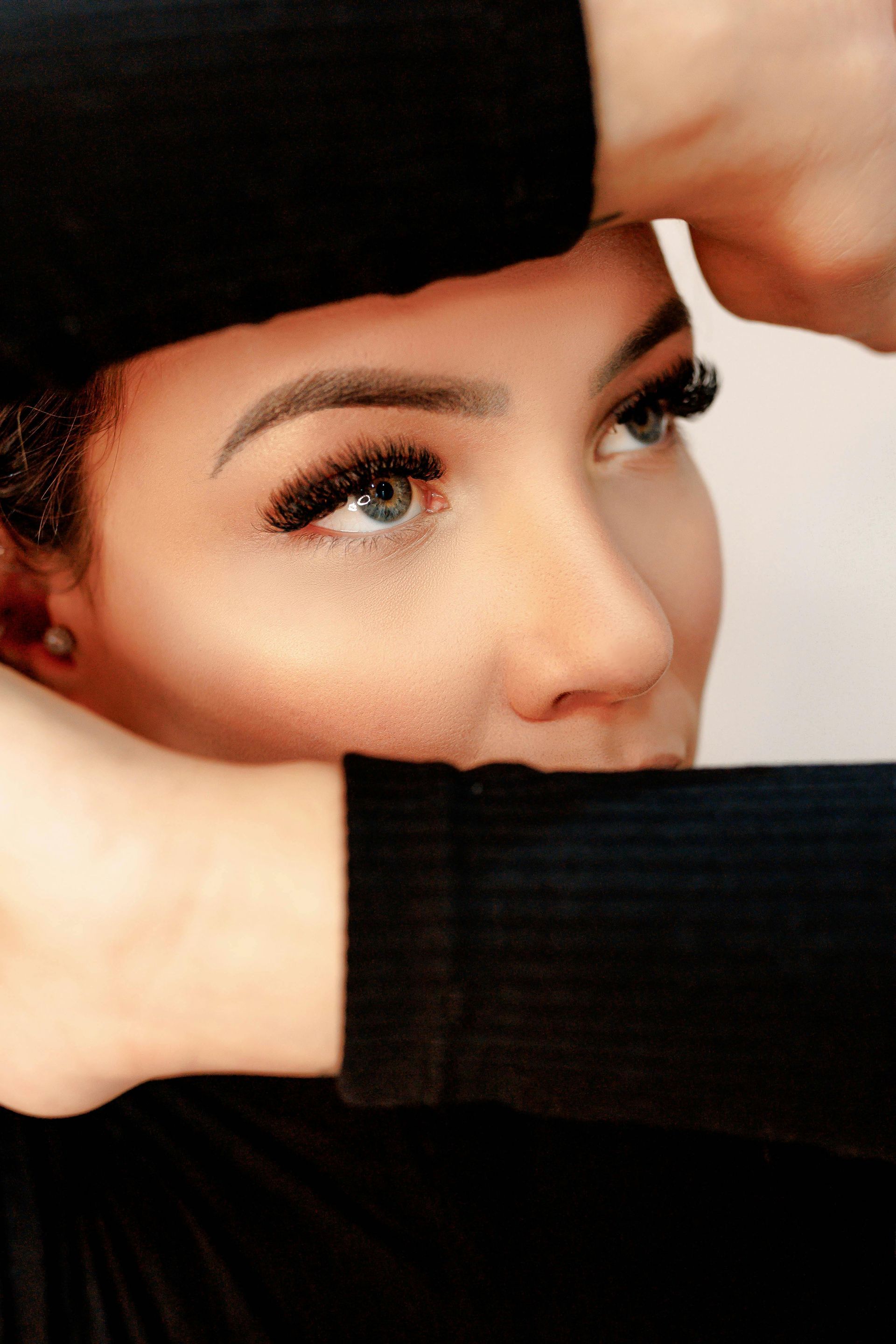 Woman with long eyelashes looking up, hands framing her face, wearing black clothing.