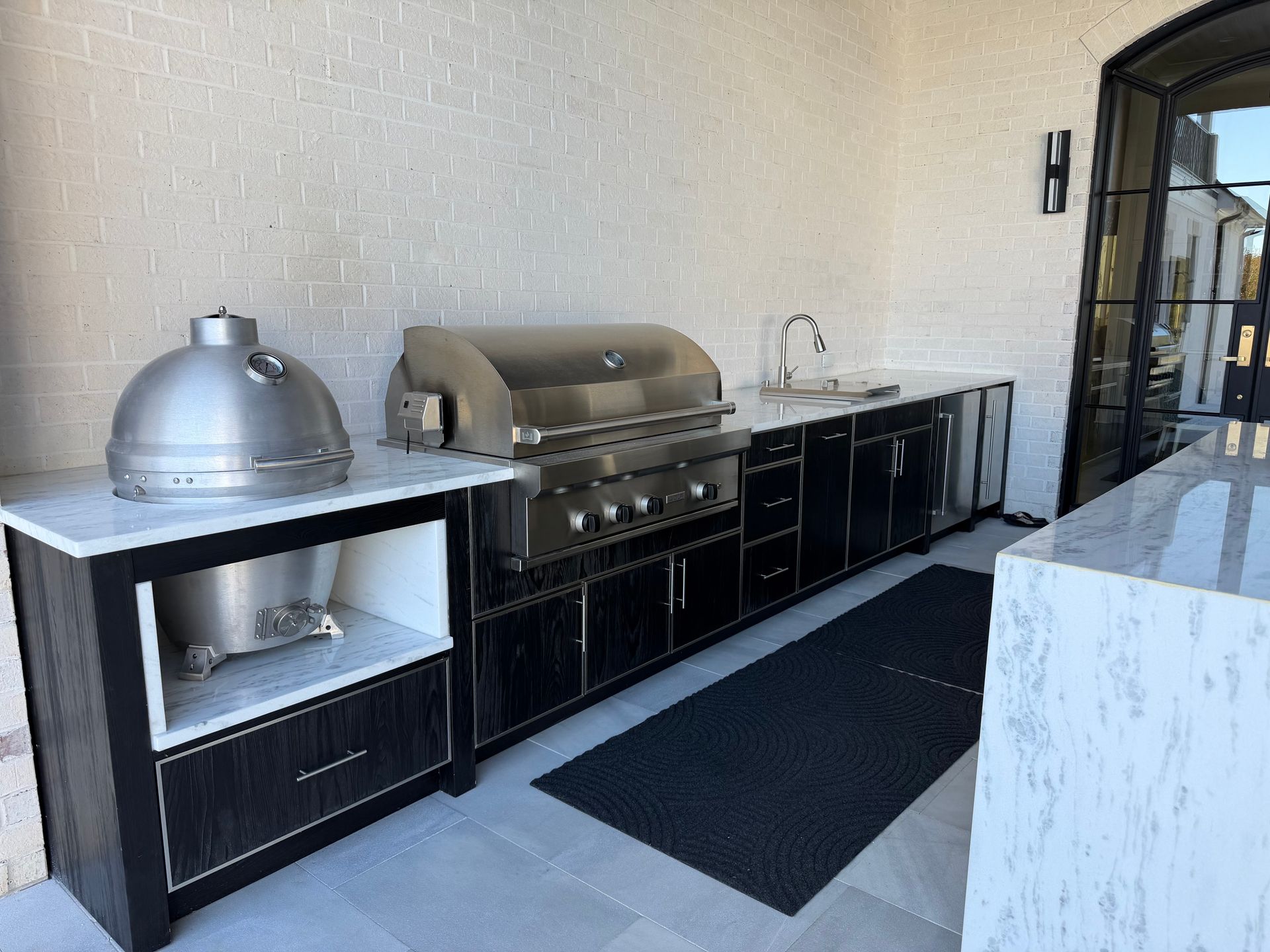 Outdoor kitchen with a grill, sink, and storage cabinets against a white brick wall.
