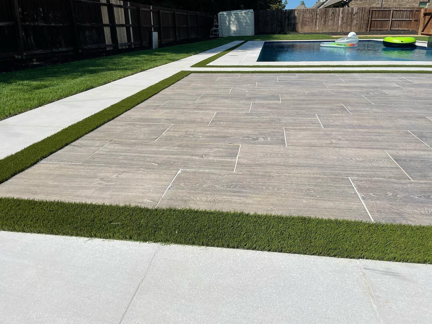 Patio with gray stone tiles, artificial turf border, and a pool in the background.