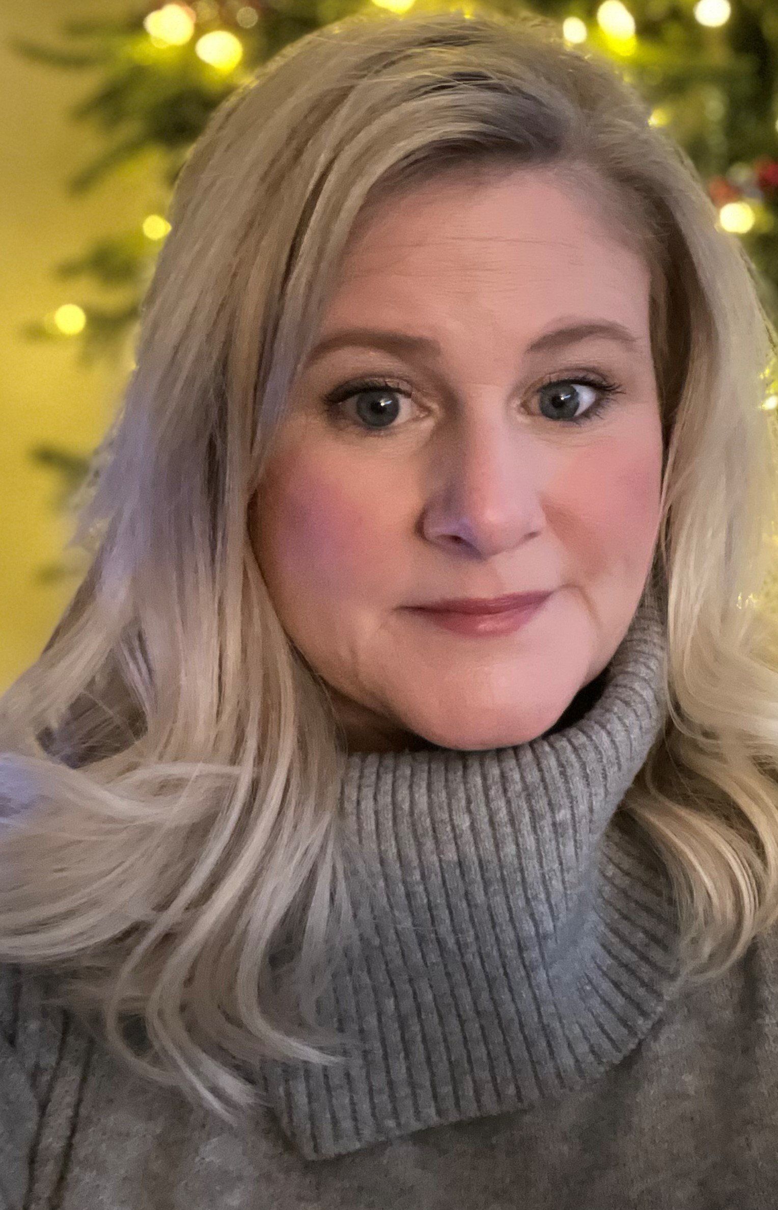 Woman in a gray turtleneck sweater, softly lit in front of blurred holiday lights.