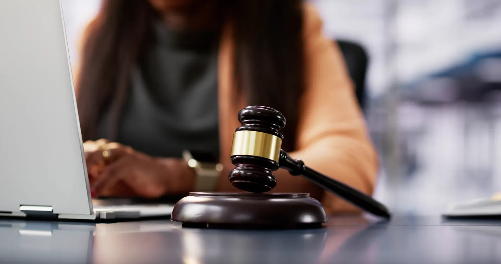 Gavel on a desk beside a laptop, with a blurred person working in the background.