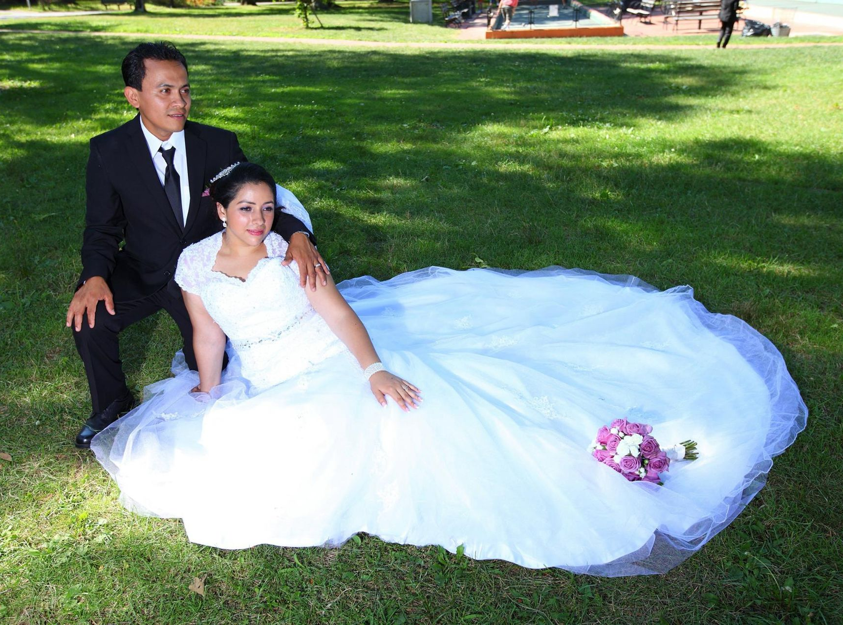 Wedding couple on grass: man in suit kneels beside woman in white gown with bouquet.