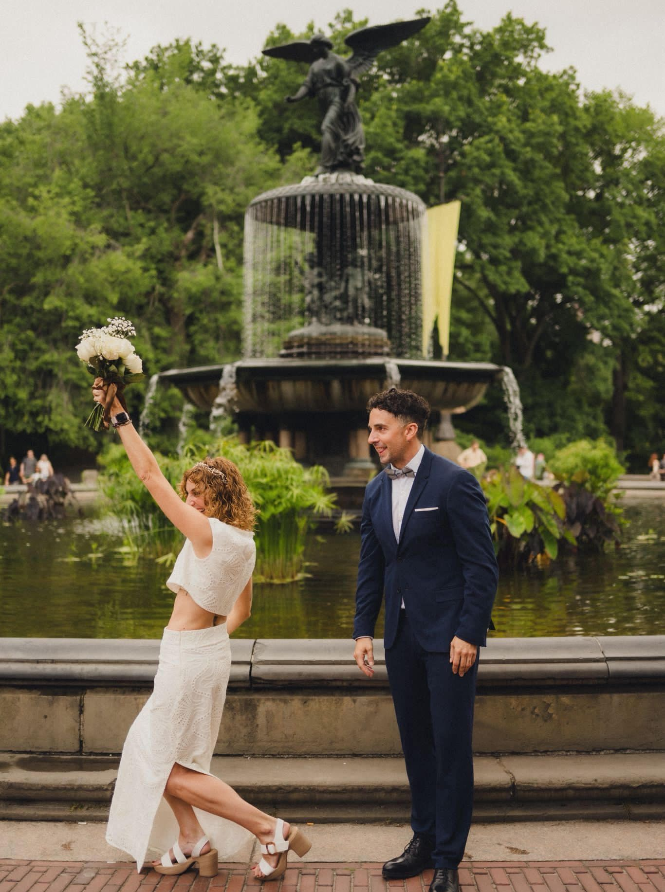 Una pareja vestida de novios posa frente a la fuente de Bethesda en Central Park; la novia sostiene un ramo en alto.