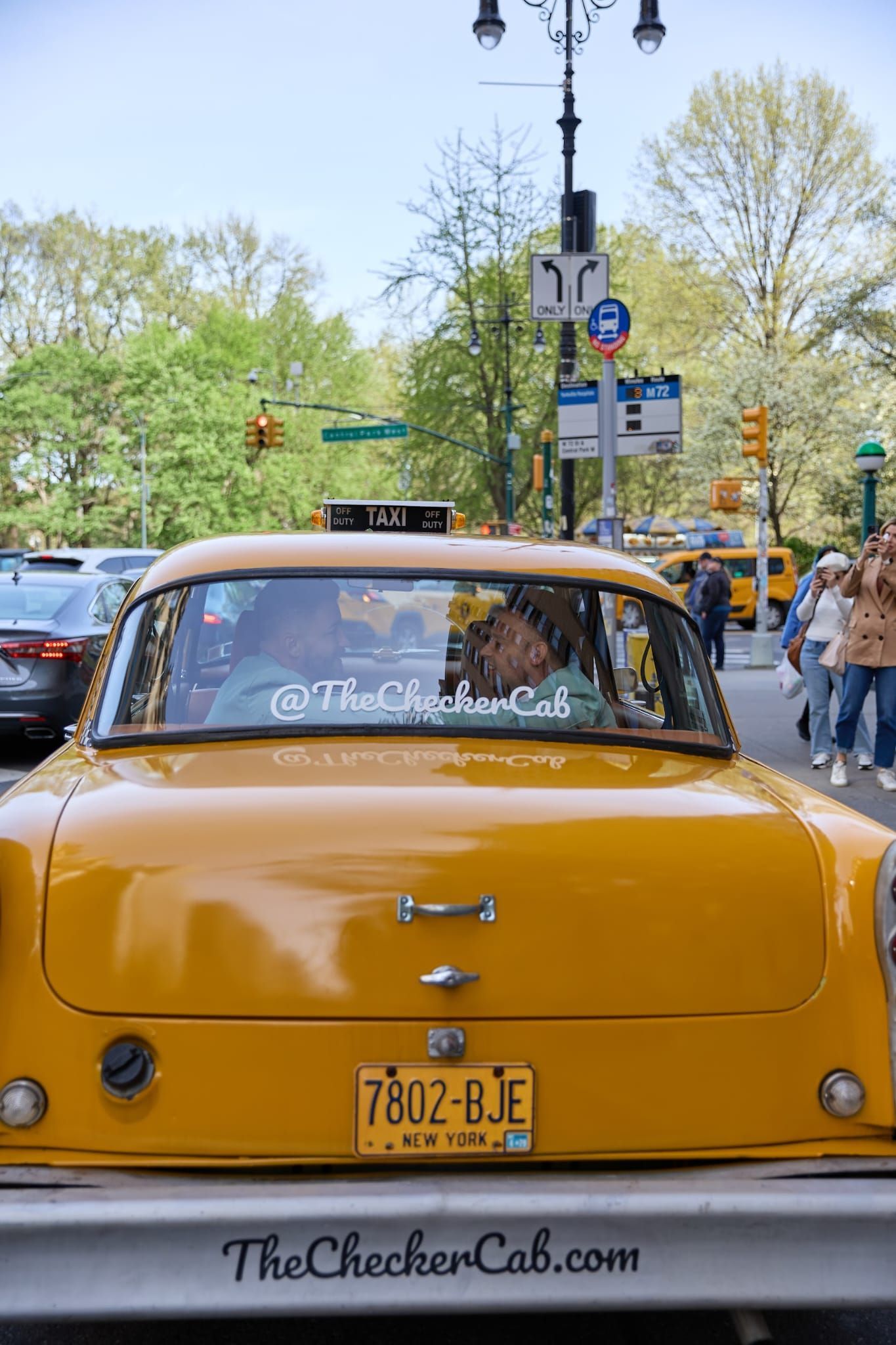 A yellow Checker cab driving down a city street, showing its license plate and the website