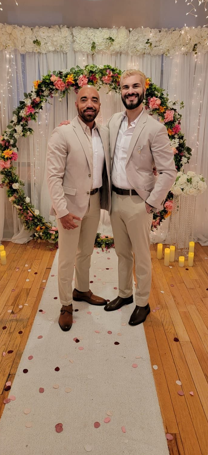 Two men in beige suits stand smiling on a white aisle runner in front of a floral wedding arch decorated with fairy lights.