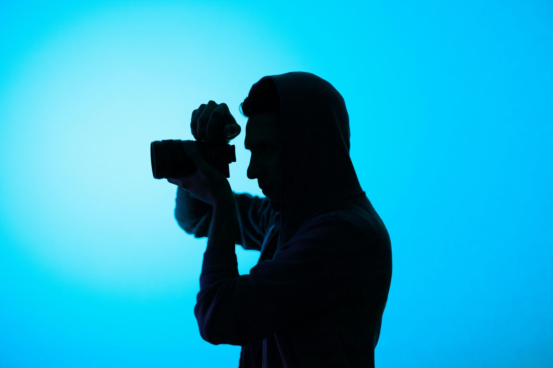 A silhouette of a person wearing a hoodie, seen in profile, holding a camera up to take a picture against a blue background.