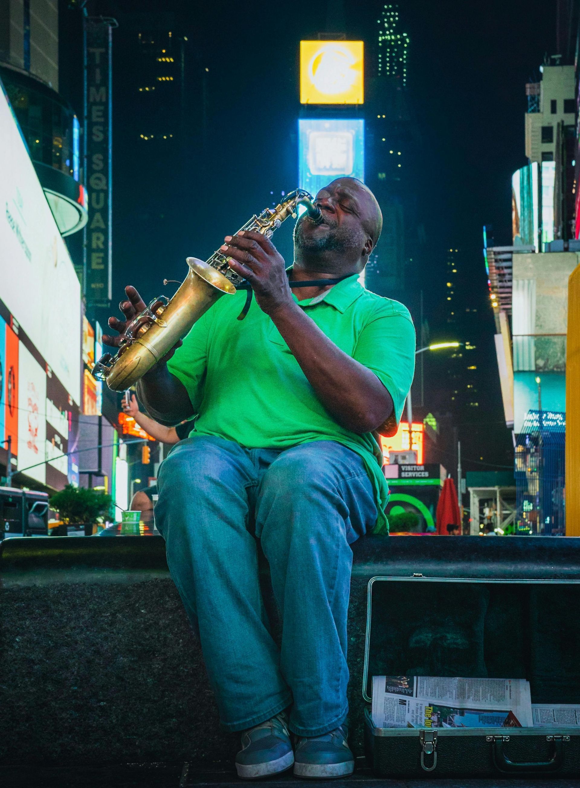 A person in a green shirt plays a saxophone while sitting in front of a case in a brightly lit Times Square at night.