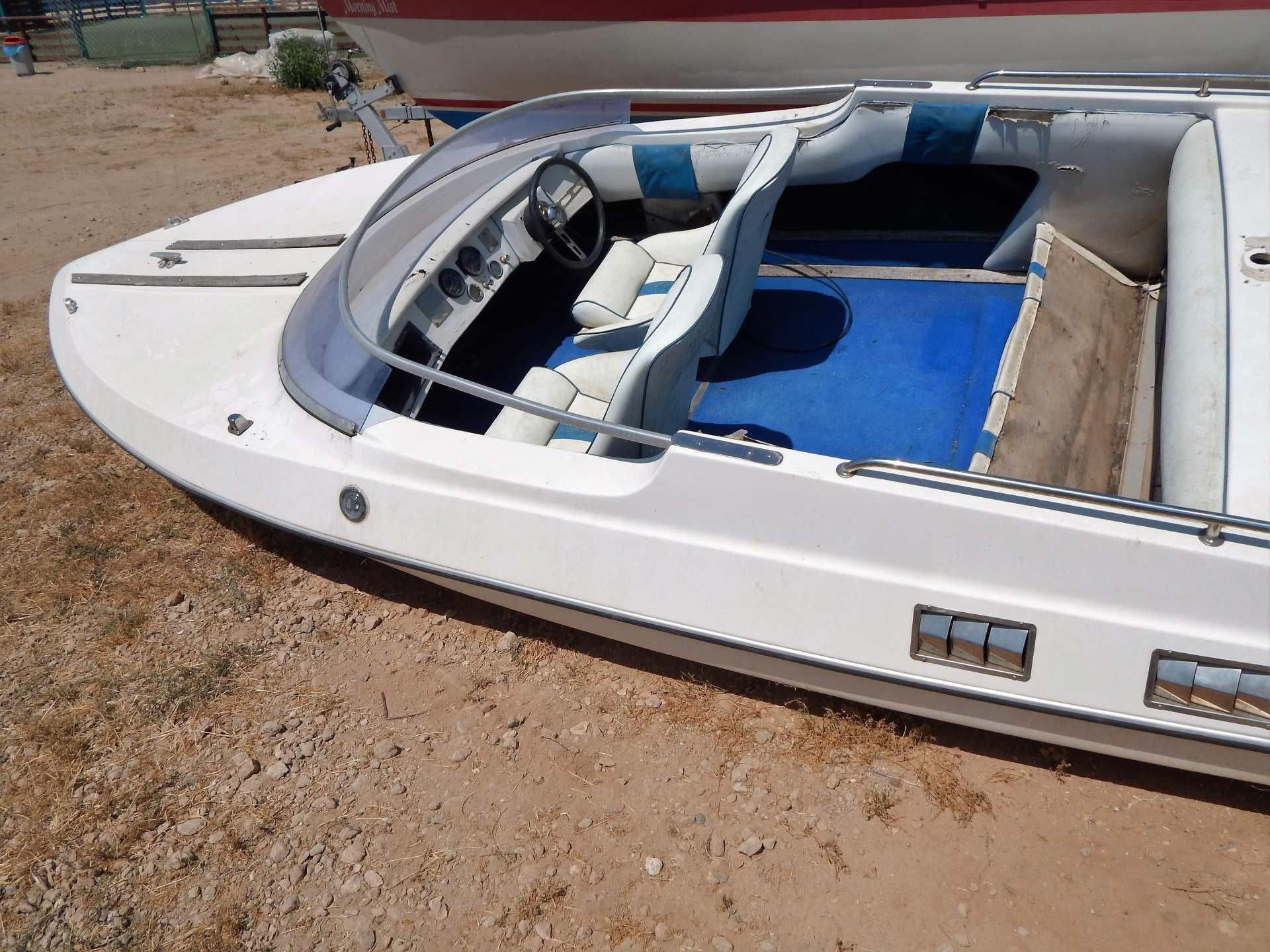 White boat with blue seats and windshield, resting on dry ground with a tan panel open on the side