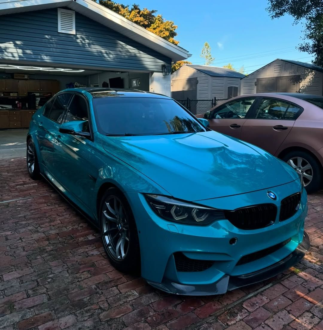 Blue BMW sports car parked in front of a house. Another car is parked nearby.