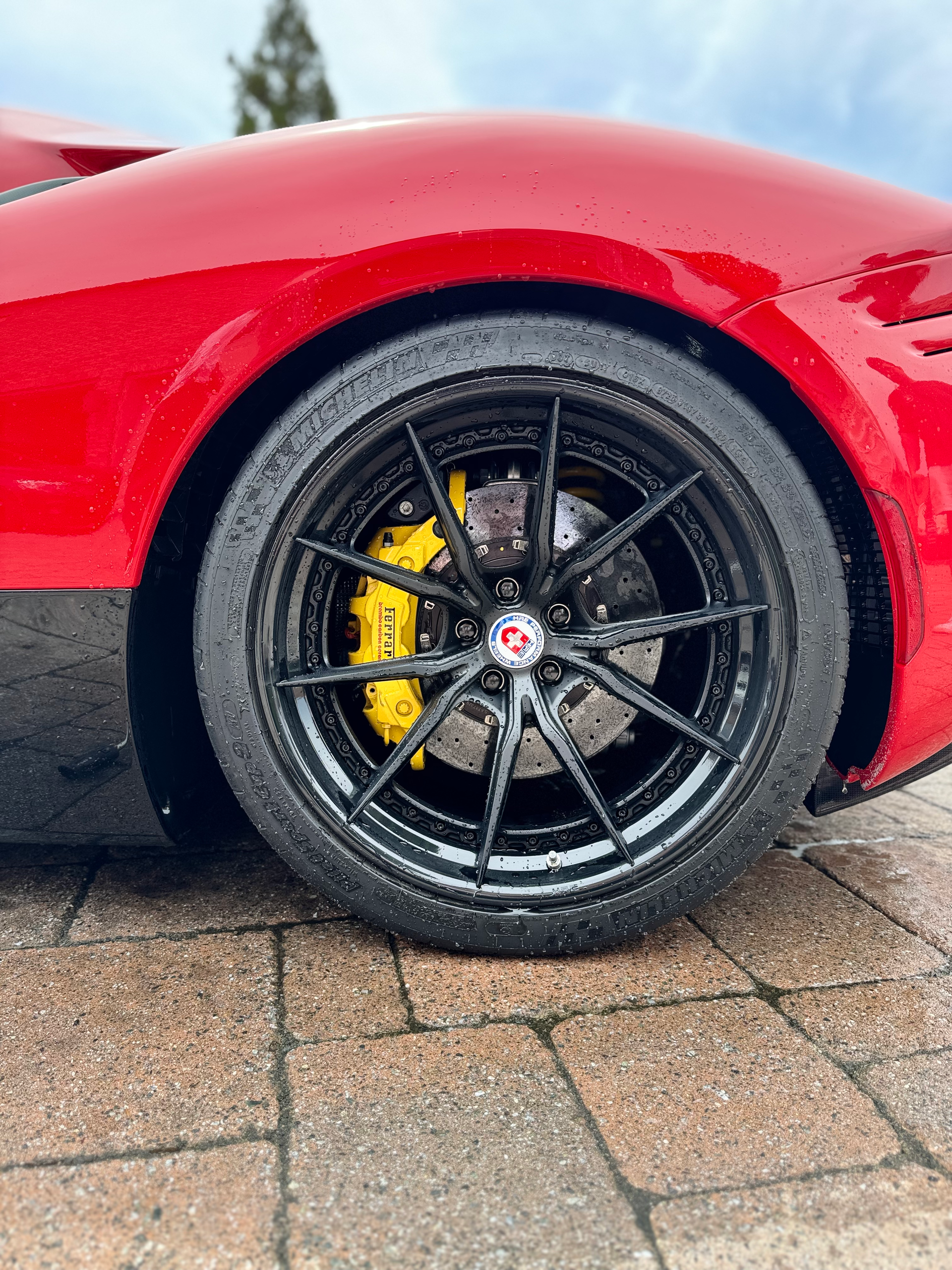 A red sports car with black wheels and yellow brake calipers is parked on a brick driveway.
