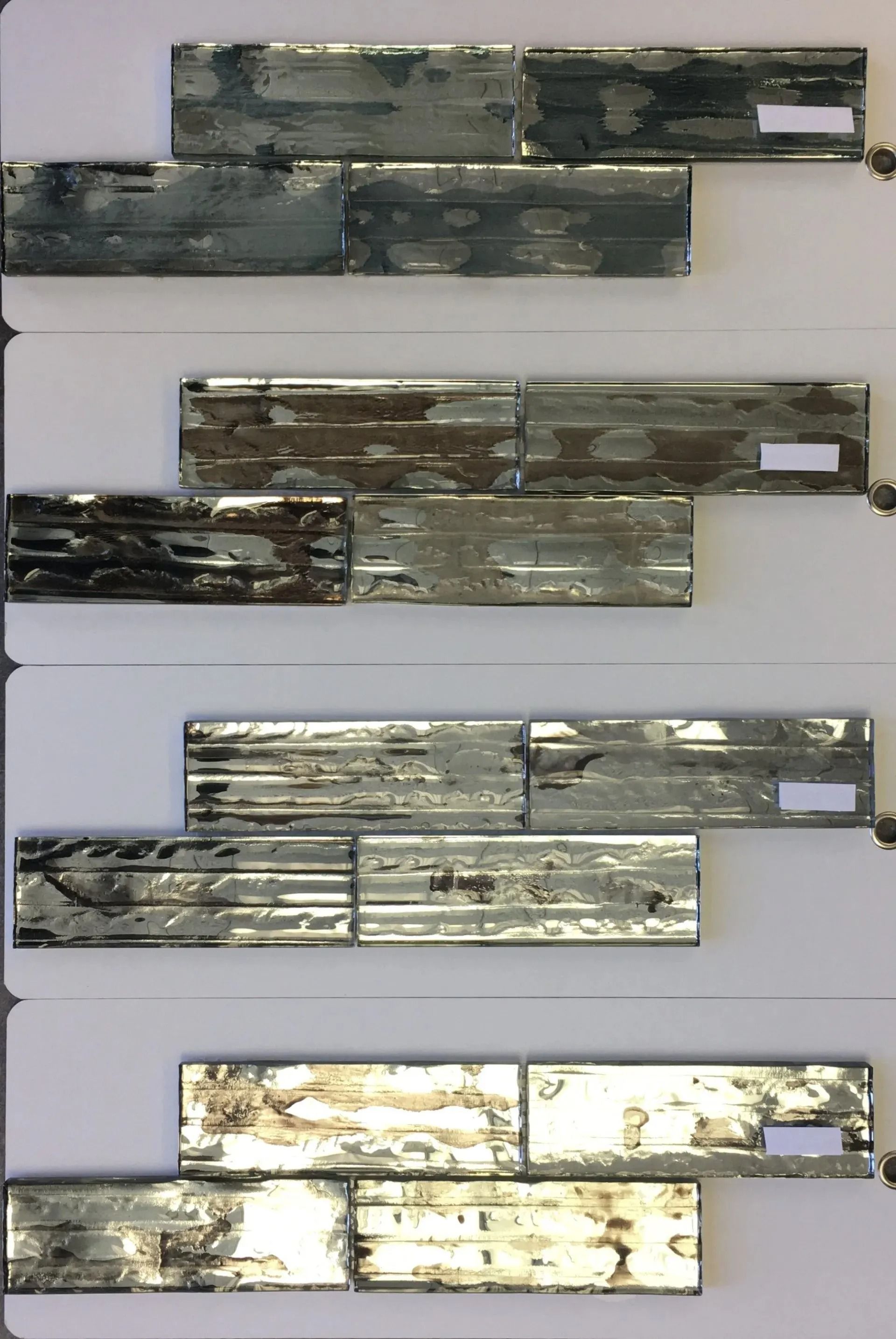 Four horizontal metal tile samples stacked vertically, each with a different reflective finish.