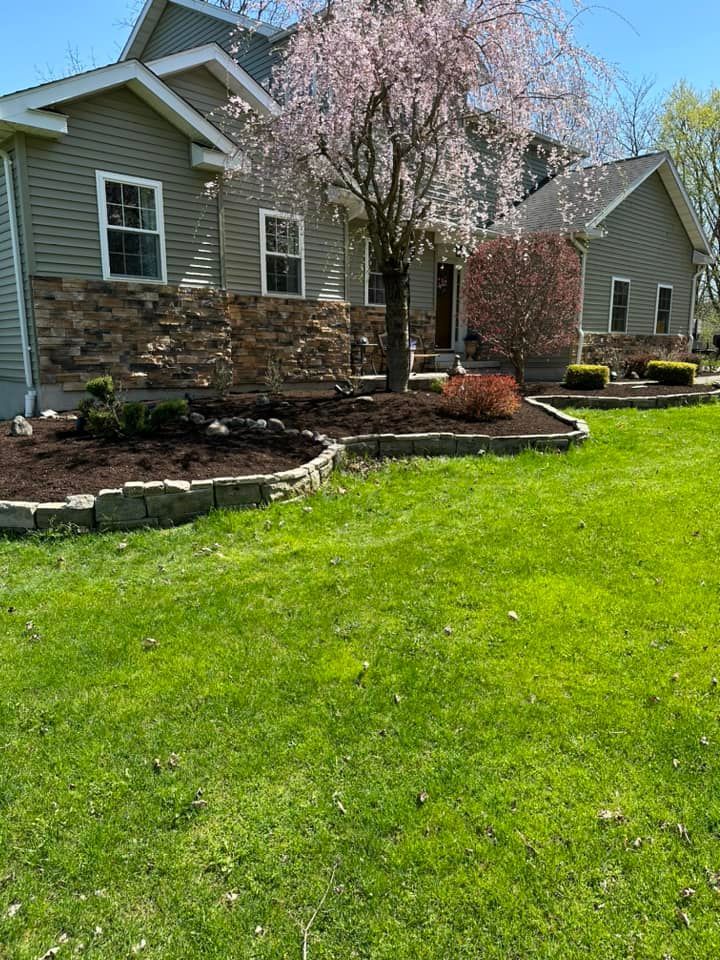 House with green siding and stone accents, a blooming tree, and flower beds on a sunny day.