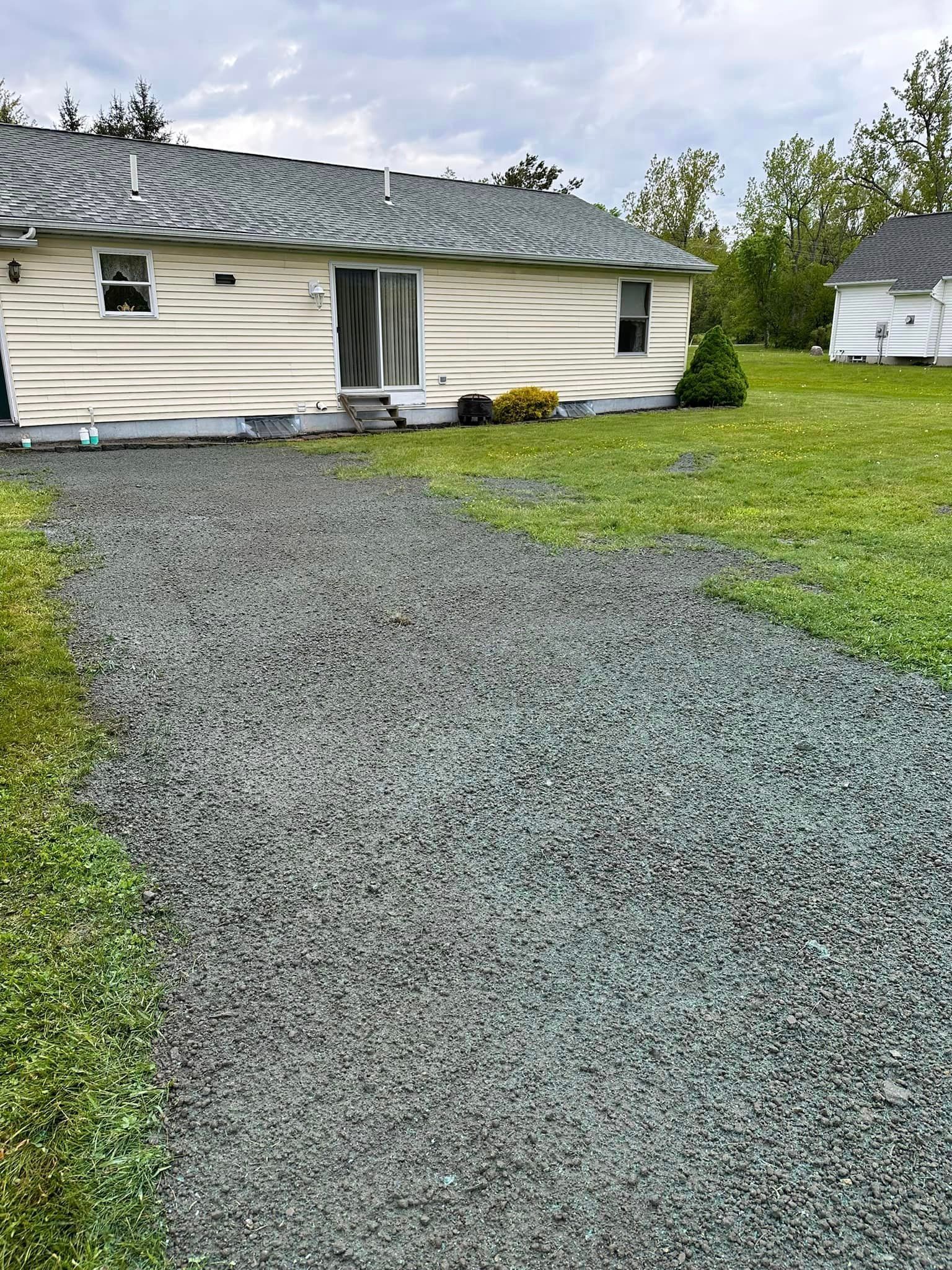 Gravel driveway leading to a one-story, light-colored building with a sliding door and a grassy lawn.