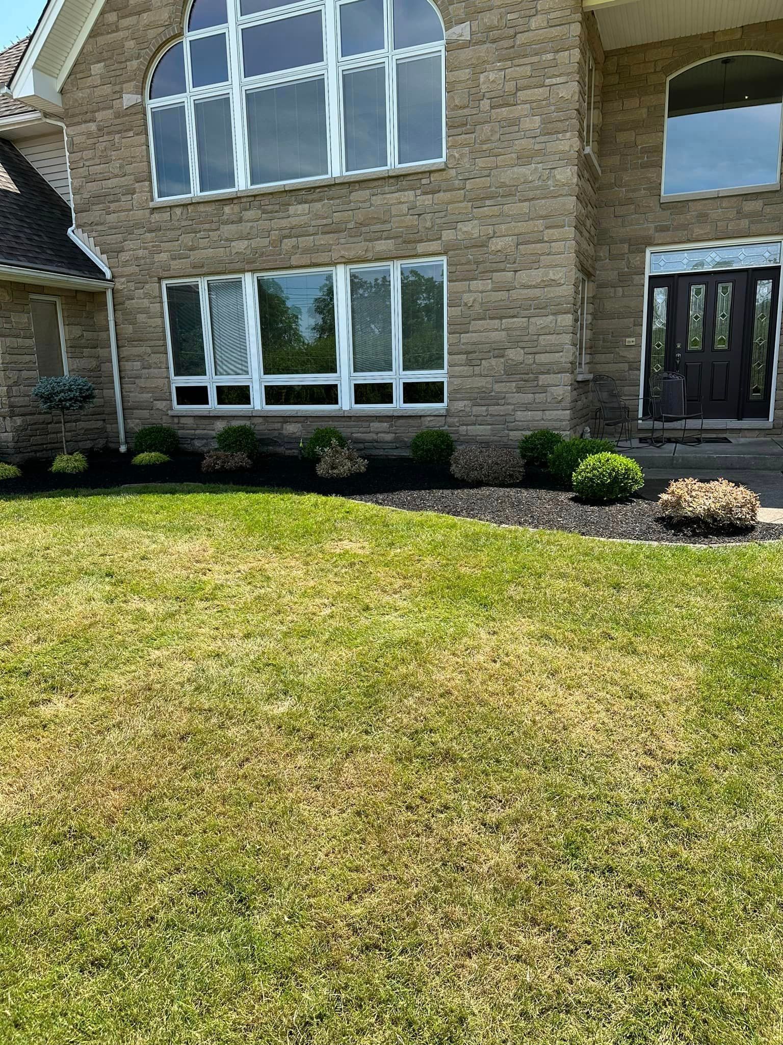 Two-story brick house with patchy lawn; black mulch beds with green shrubs in front.