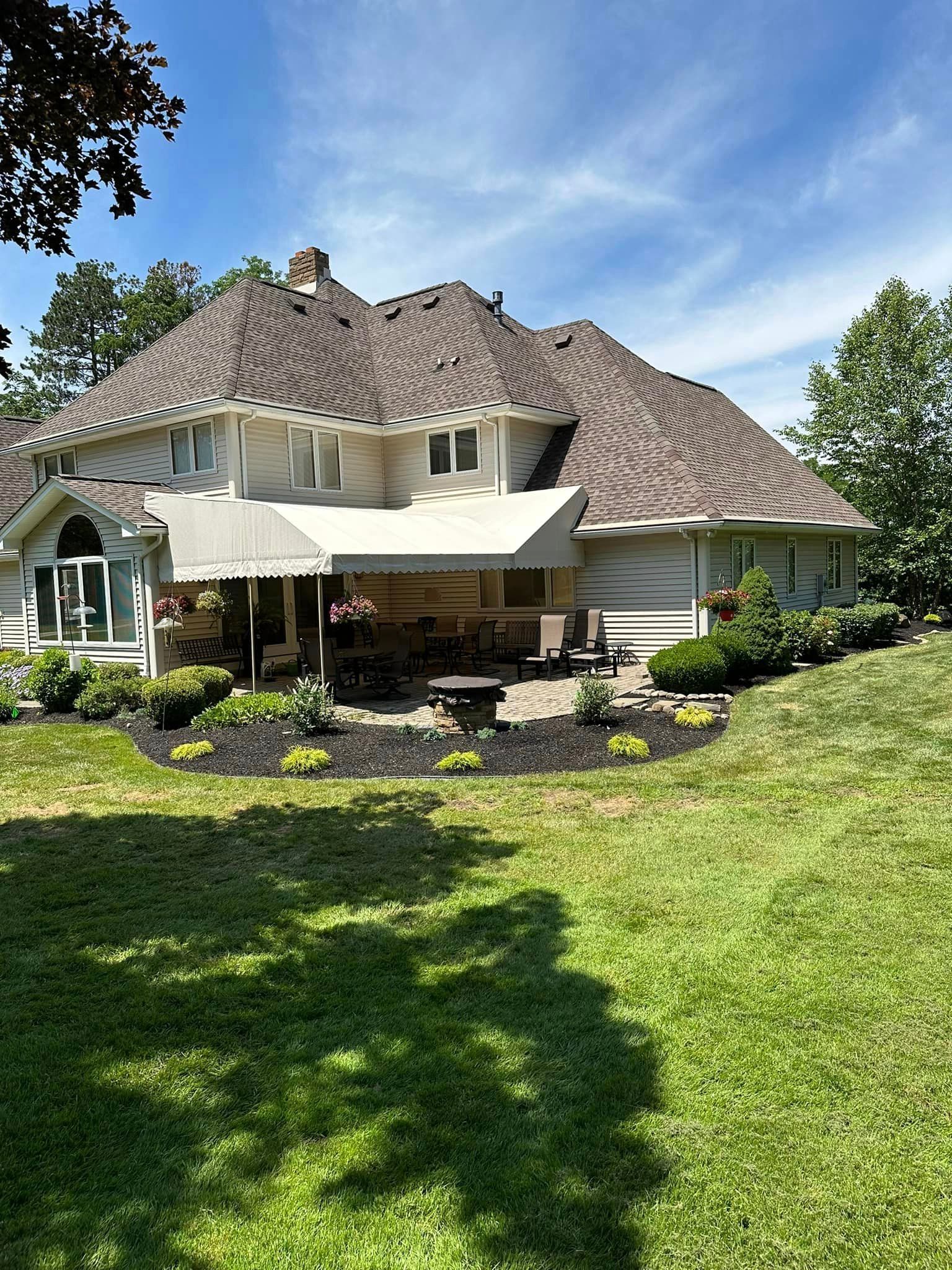 Back of a beige house with a brown roof and a patio with awnings and landscaping.