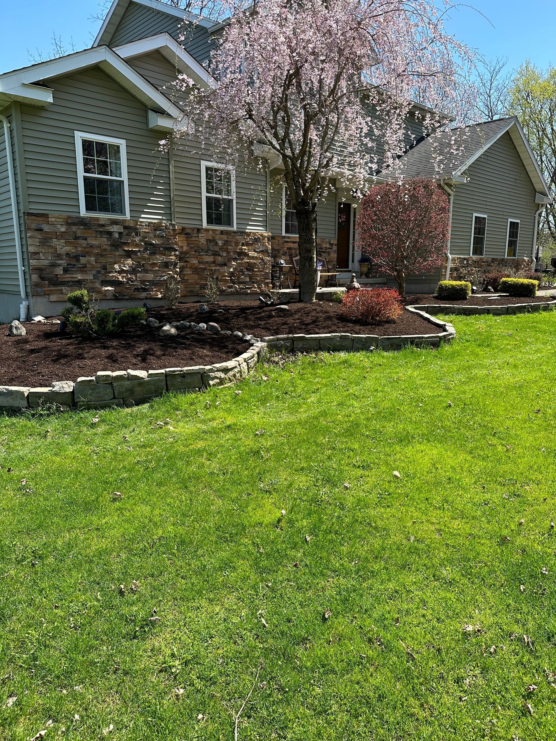 House with green siding and stone accents, front yard with flowering tree and garden.