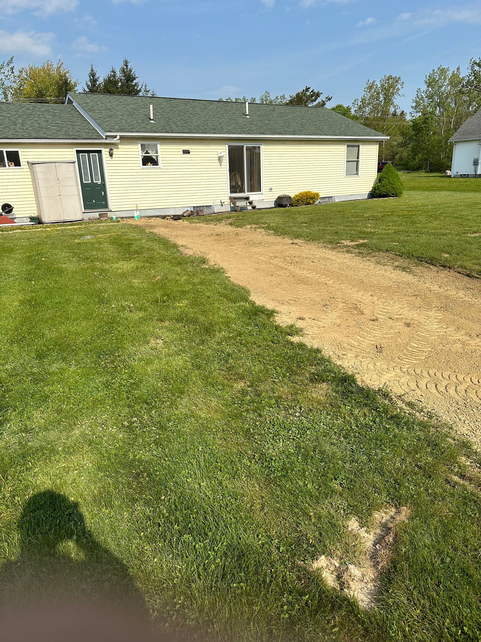 A small, light-yellow house with a gravel driveway. Green grass surrounds the driveway under a blue sky.