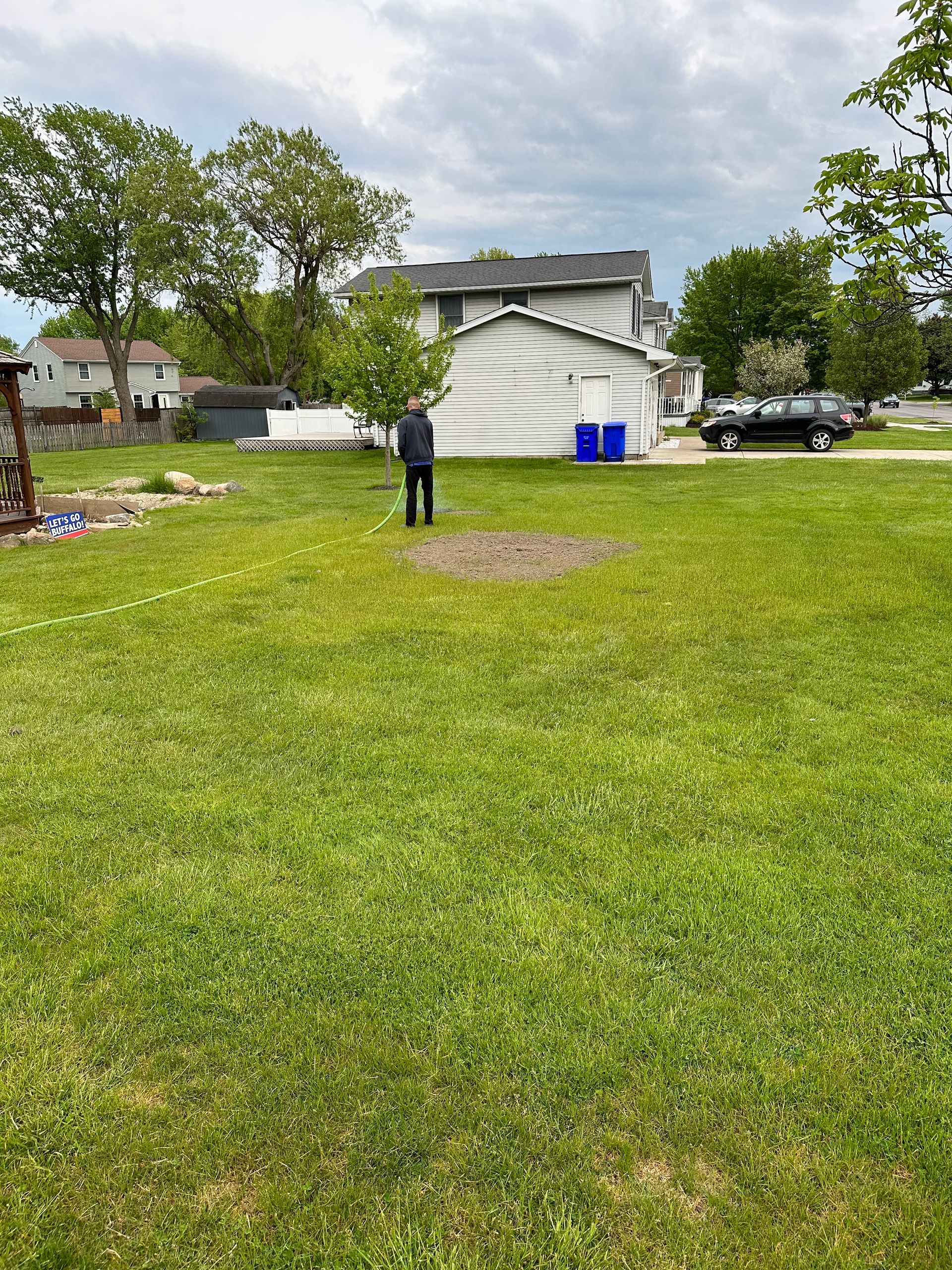 Person raking in grassy yard; shed and houses in background. Overcast sky.
