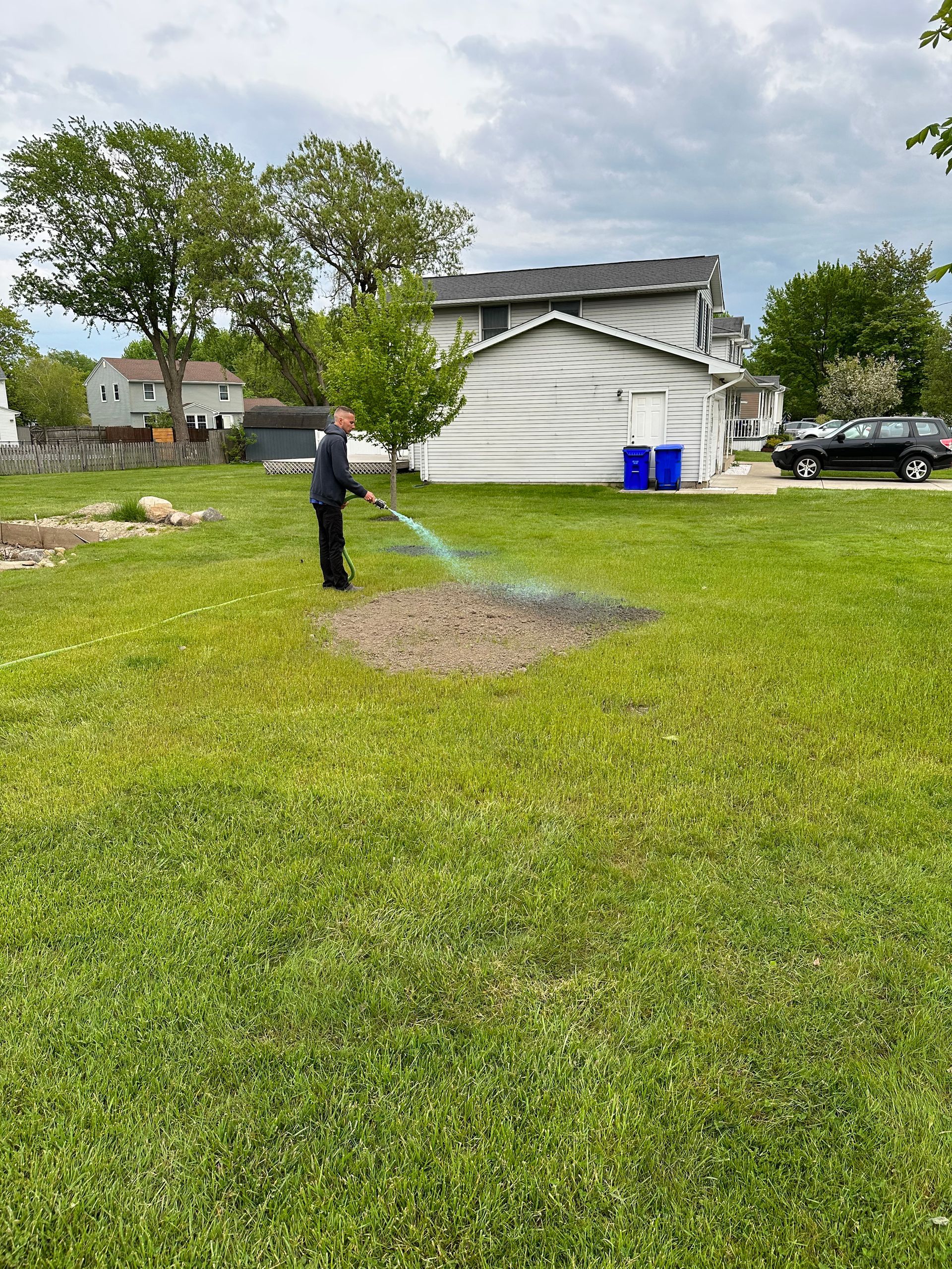 Person waters a patch of bare earth in a grassy yard, aiming the hose.