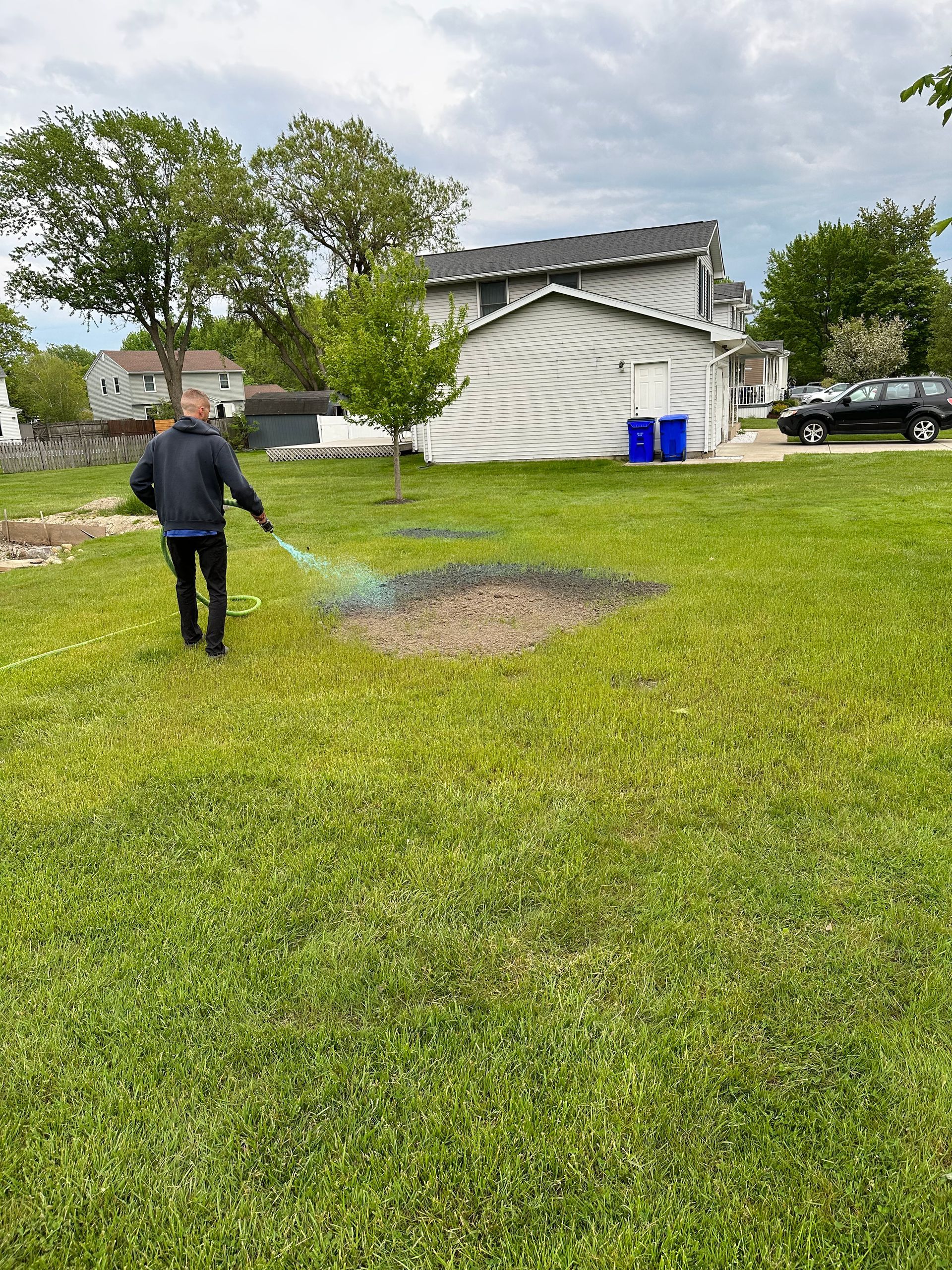 Man waters a patch of bare earth in a grassy yard, with a house and shed in the background.