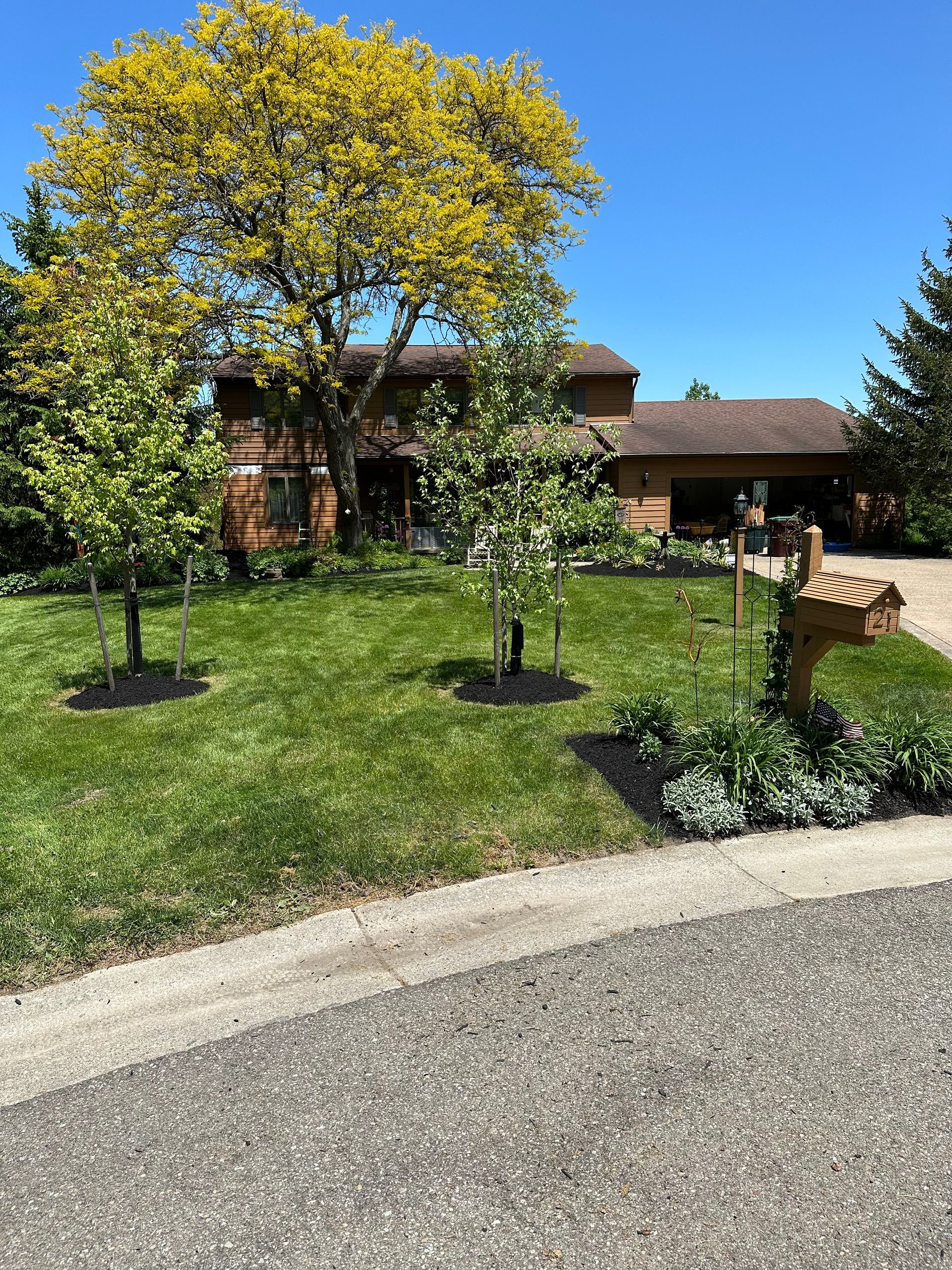 A two-story brown house with green lawn, trees, and a wooden mailbox on a sunny day.