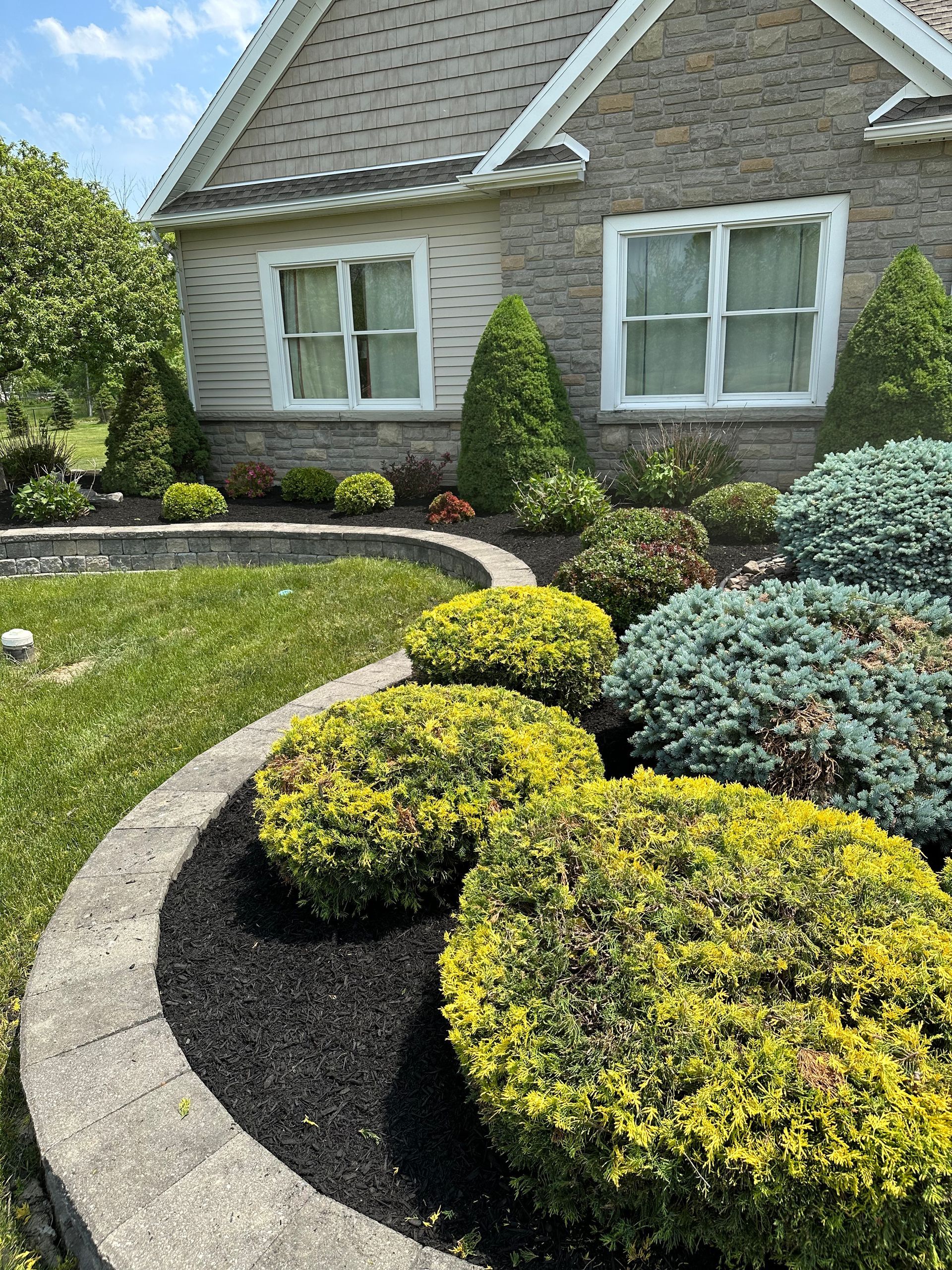 Lush landscaping with bushes, a curved retaining wall, and a house with two windows in the background.
