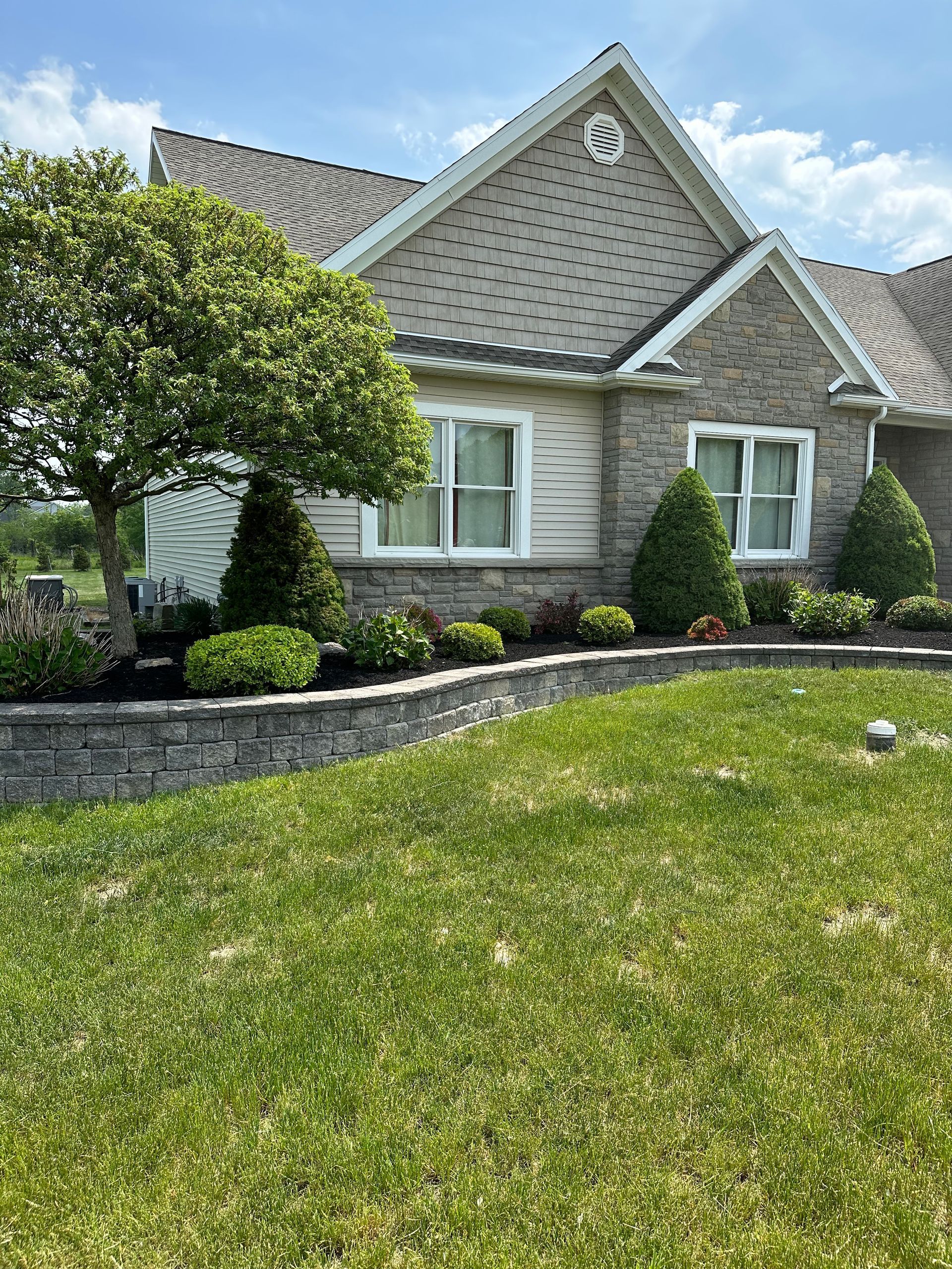 Landscaped front yard with retaining wall, house with beige siding, green grass, blue sky.
