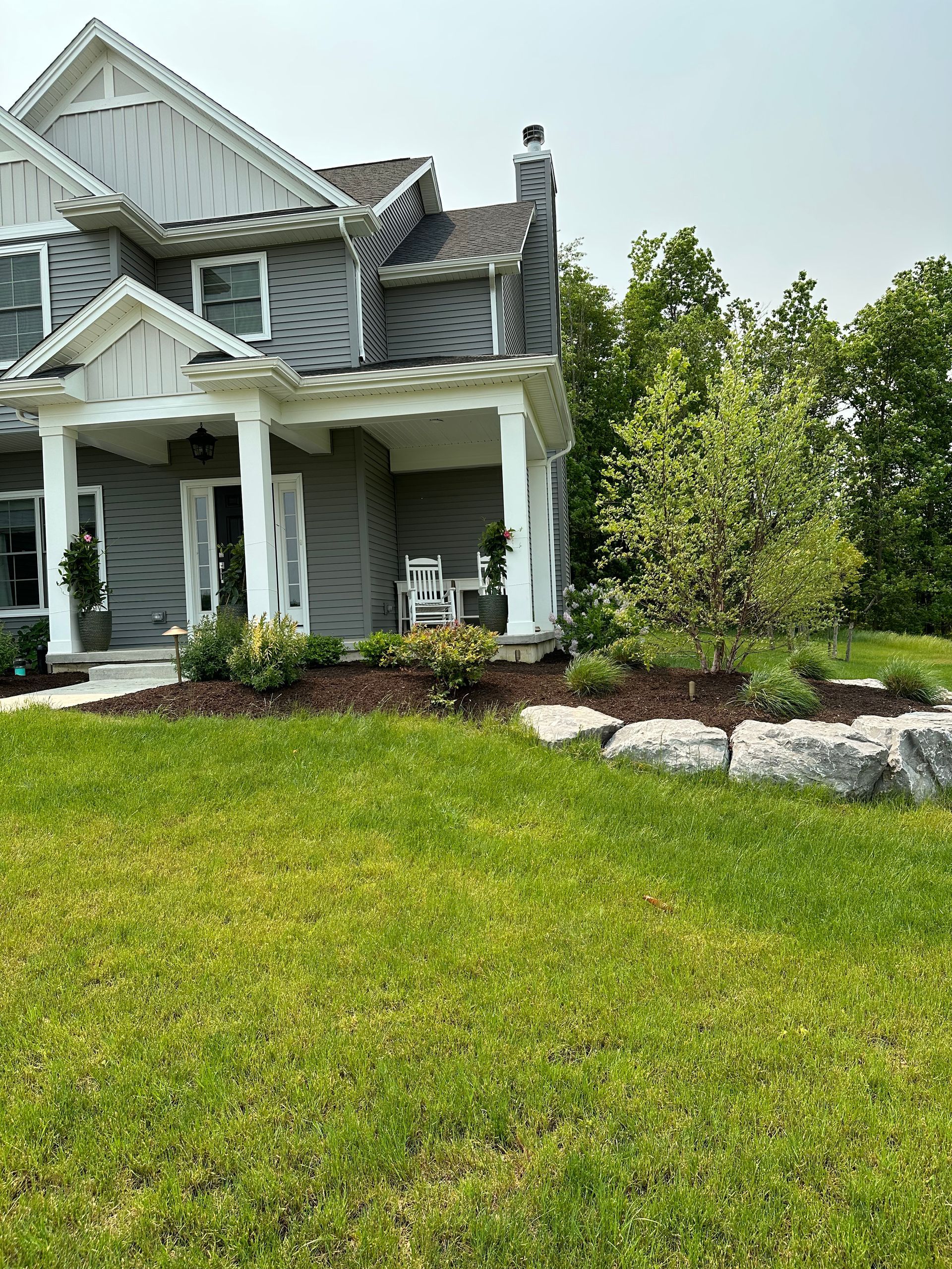 Grey house with white trim, porch, and landscaped yard. Green grass and trees in background.