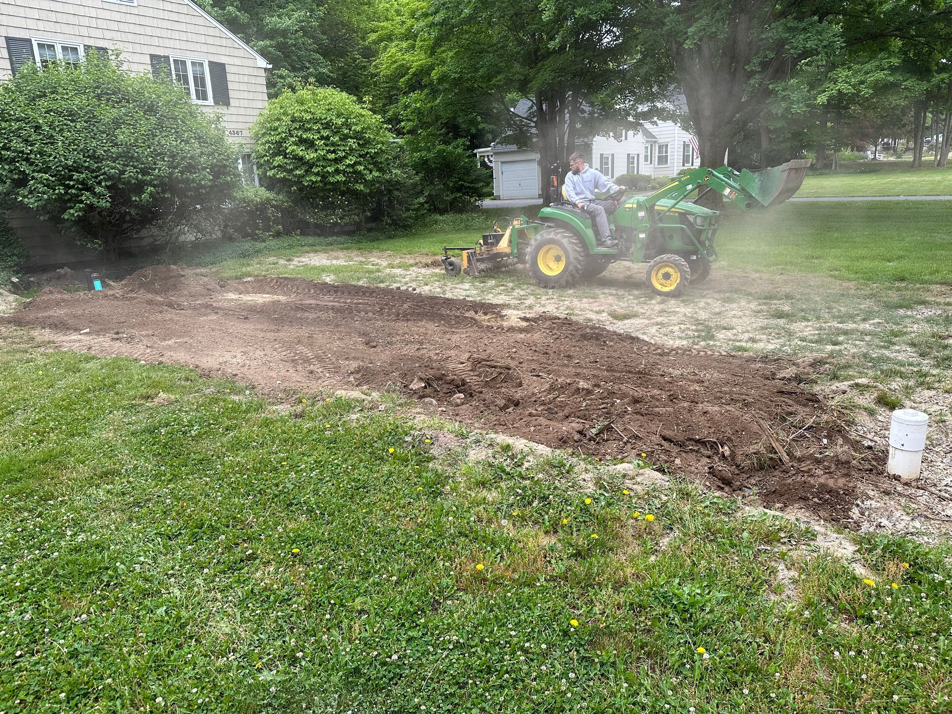 A tractor tilling soil in a yard. A person is driving the tractor in a grassy area.