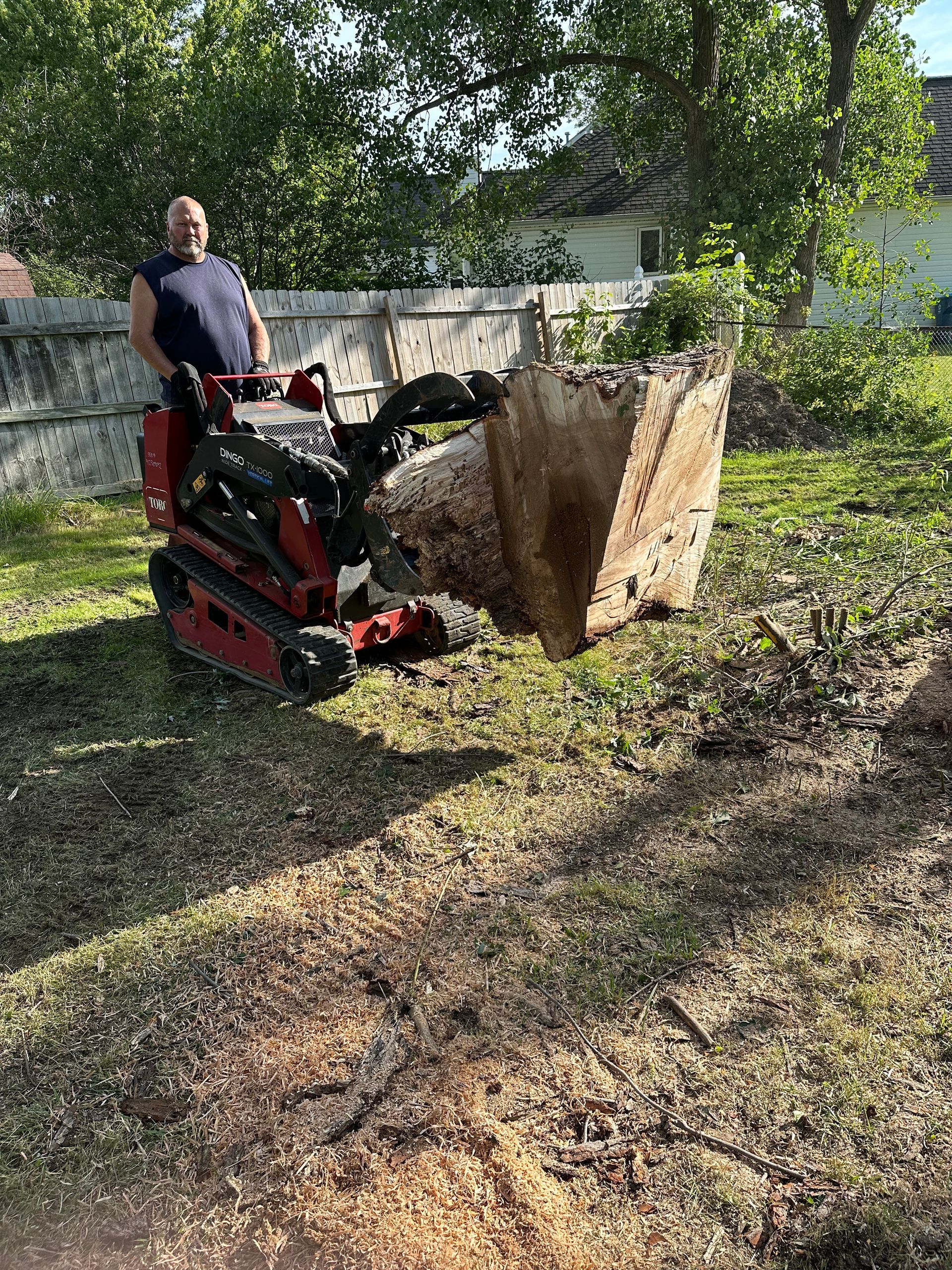 Man operating a red mini-excavator with a bucket full of dirt in a yard with a fence and trees in the background.