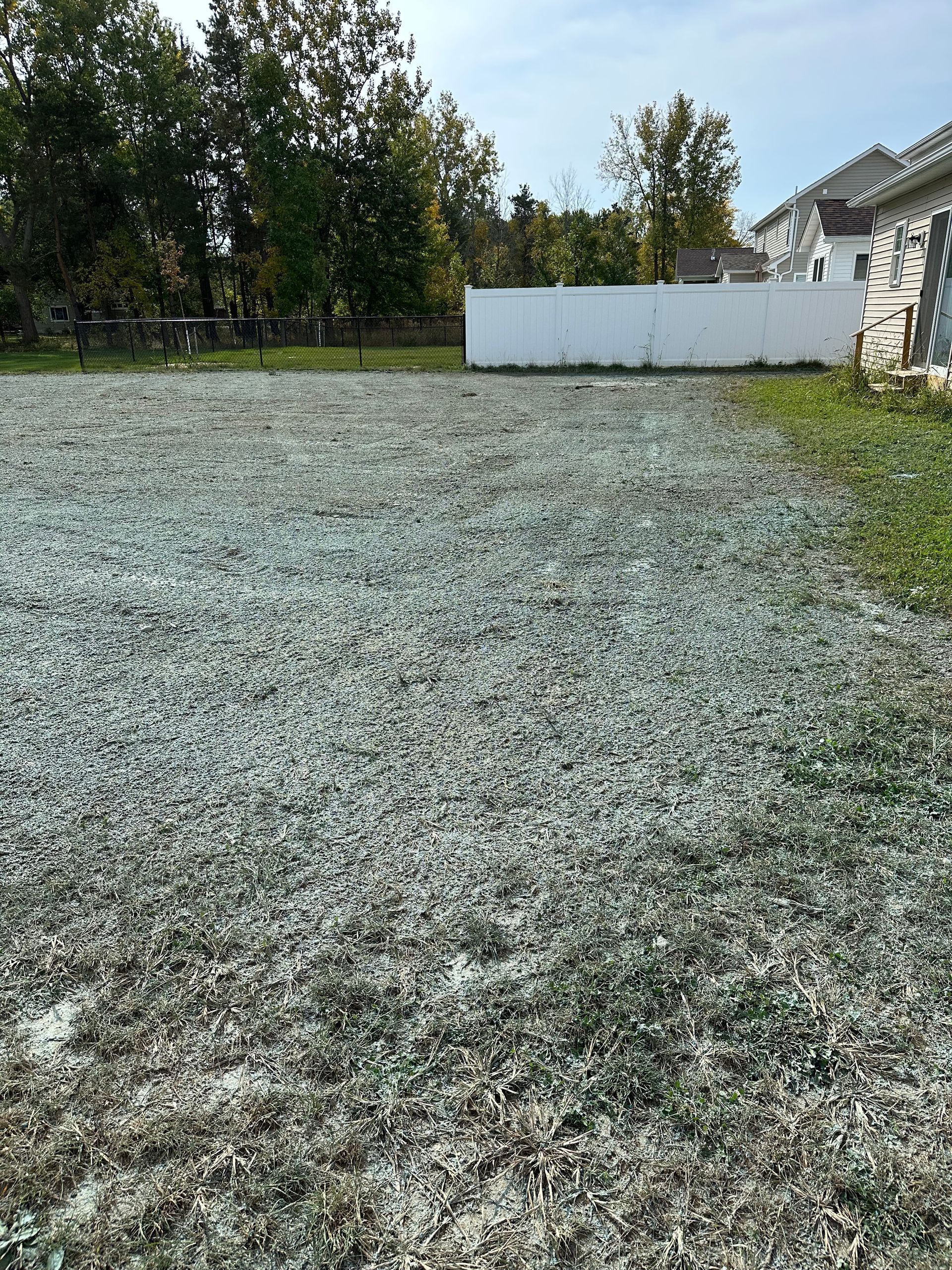 Gravel-covered lot next to a white fence and house with green trees in the background.