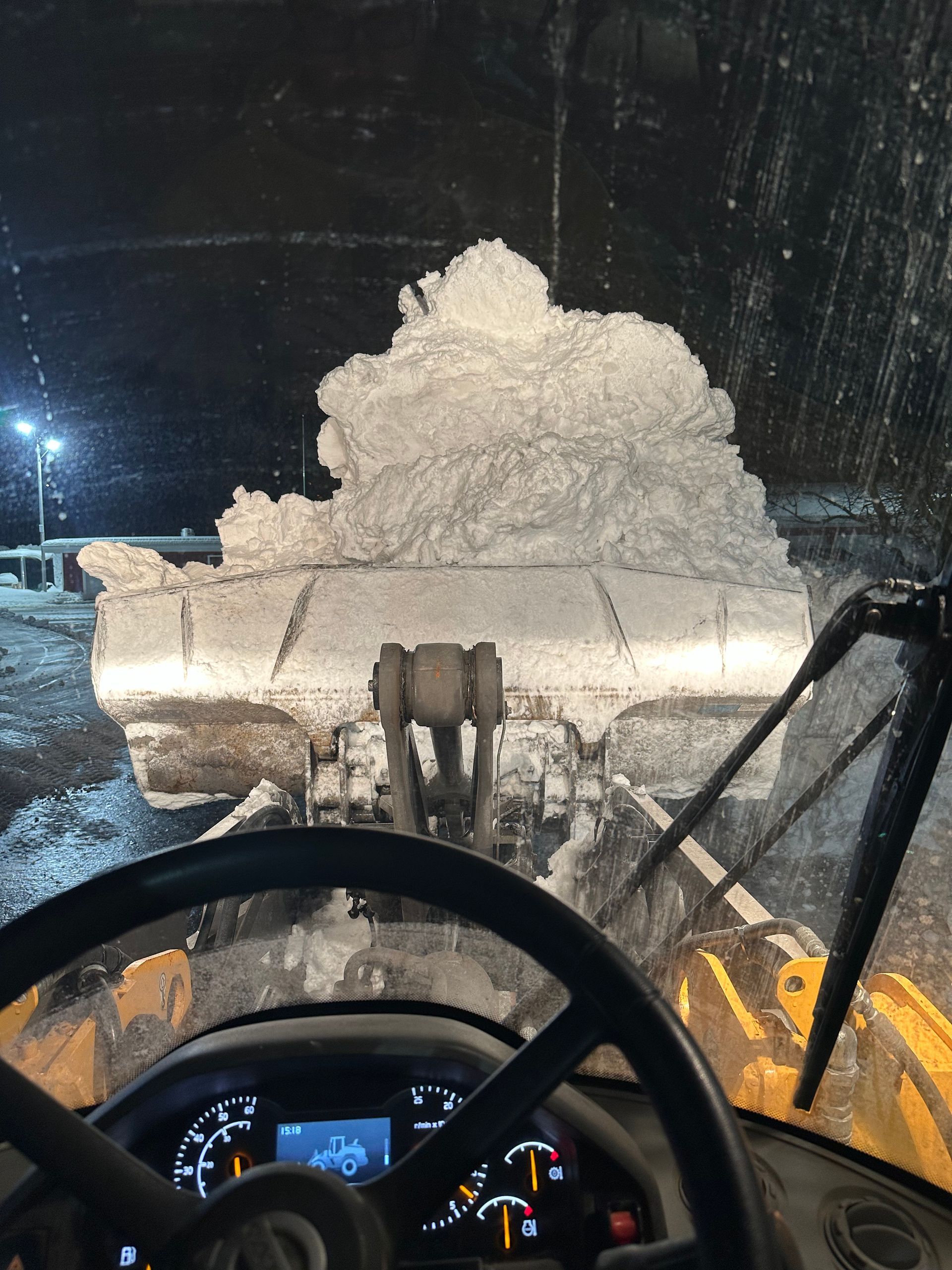 Snowplow clearing snow at night; view from inside the cab with steering wheel and lights visible.