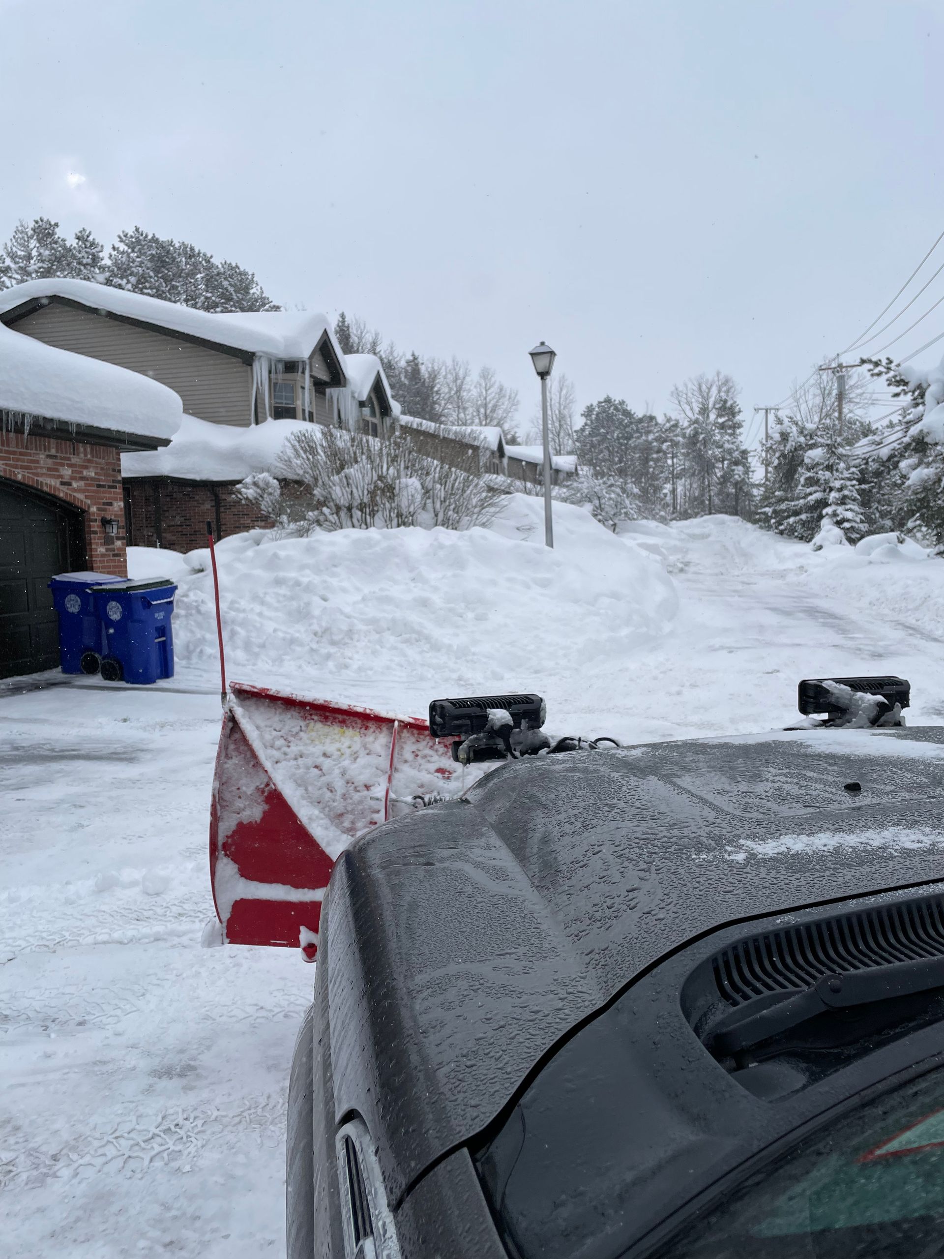 A truck with a snowplow clearing a snow-covered driveway in front of a house.