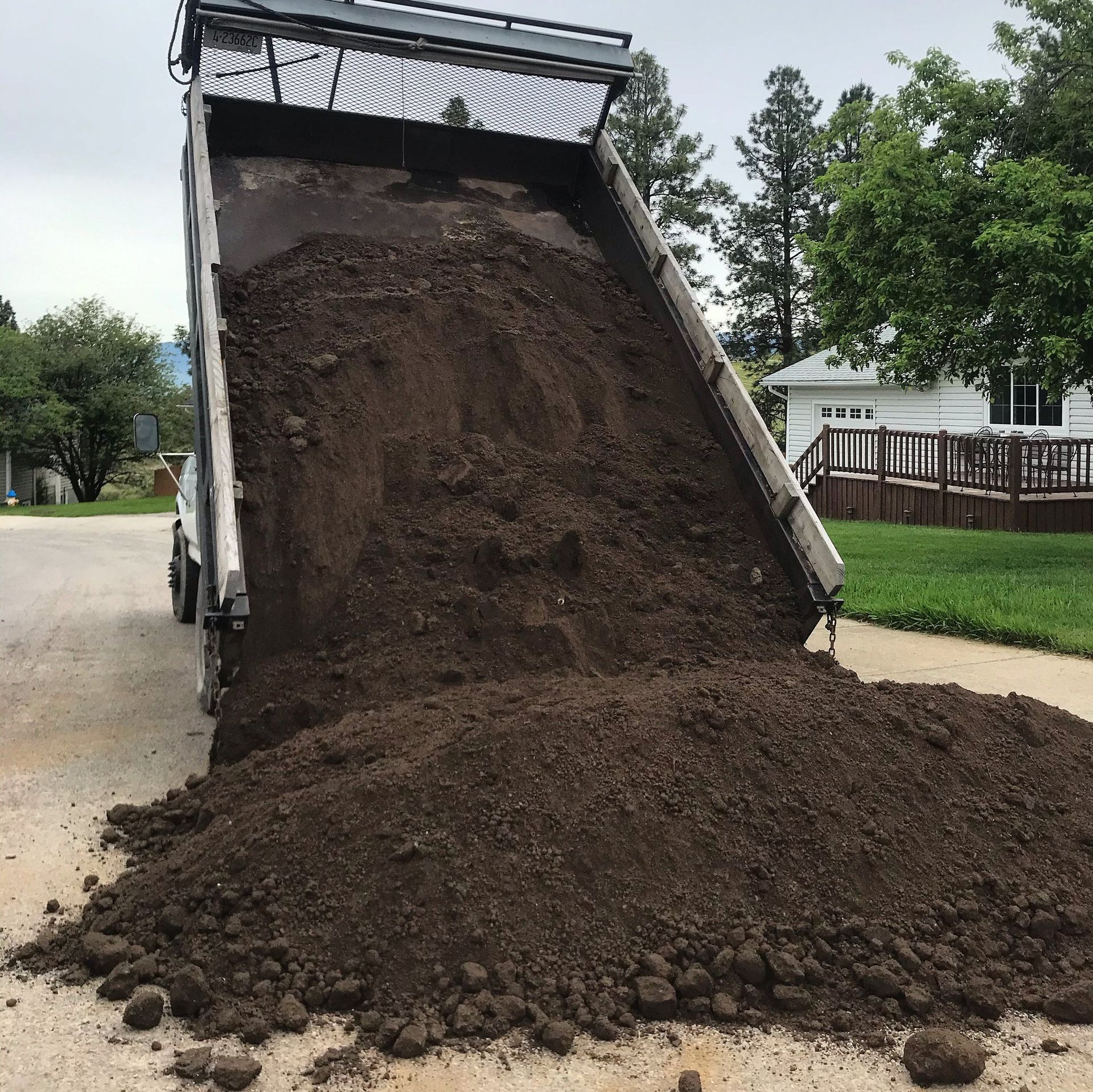 Dump truck dumping a pile of dark soil onto a paved road in a residential area.