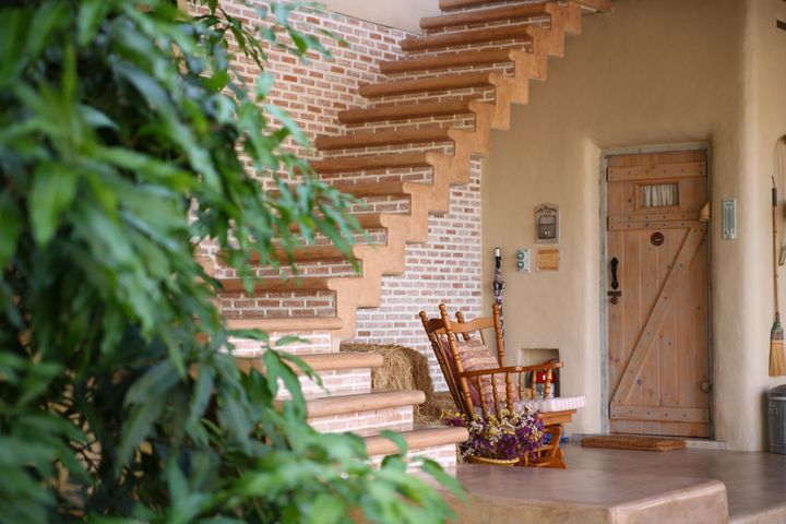 Porch with wooden rocking chair, brick stairs, and a rustic door beside lush greenery
