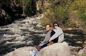 A man and a woman are sitting on a rock near a river.