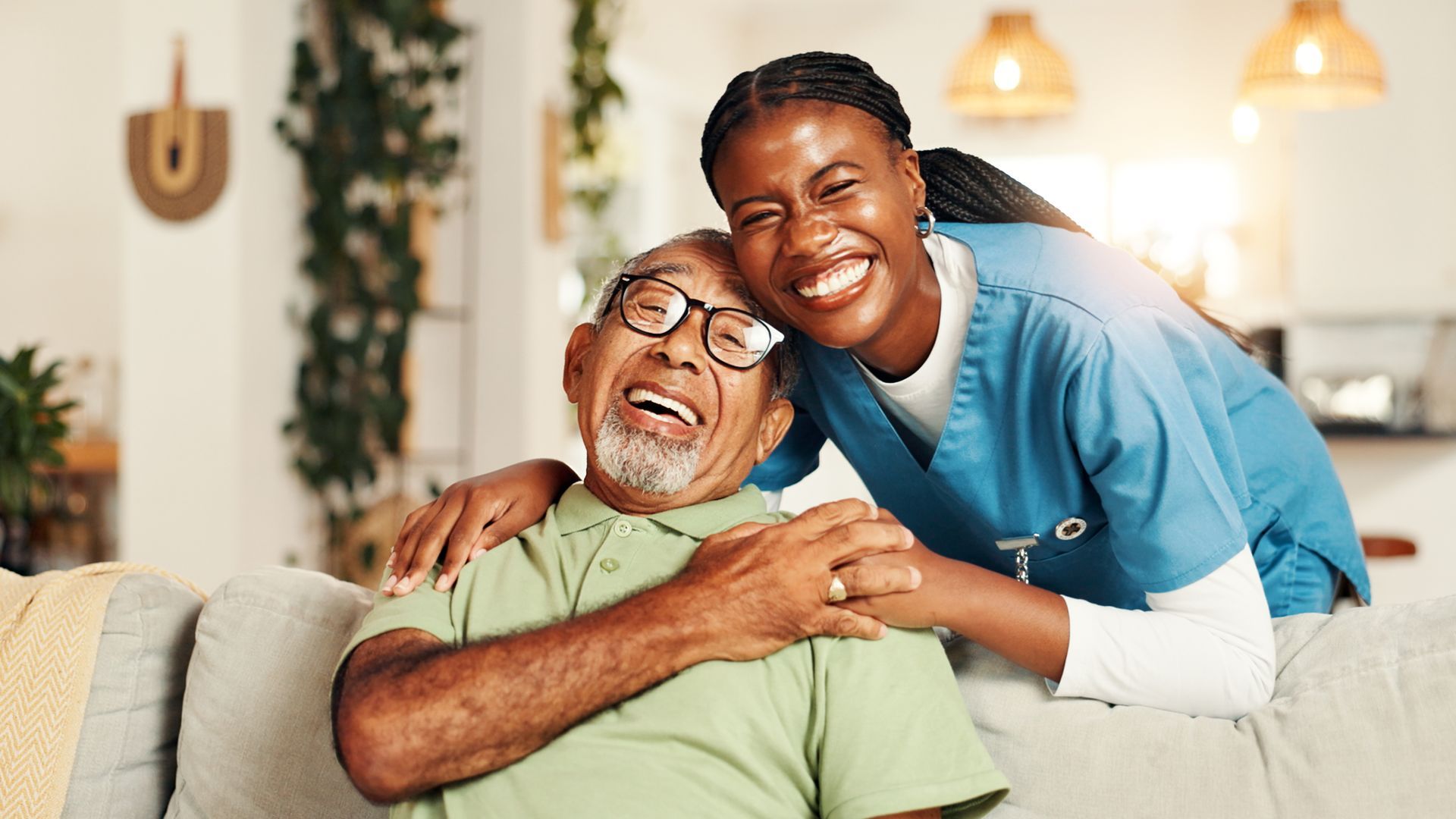 A caregiver embraces and holds the hand of an older person, both smiling gently while sitting on a leather sofa.