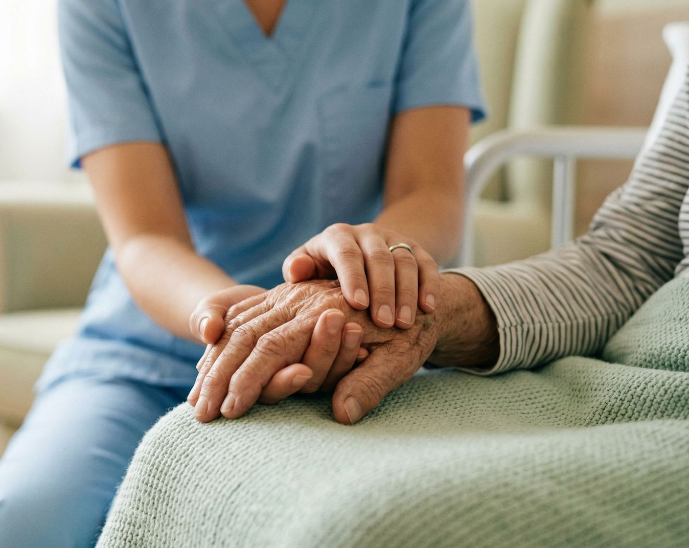 A person in blue scrubs holds the wrinkled hand of another person, providing comfort and support.