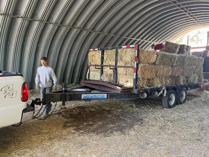 A person standing next to a truck pulling a trailer loaded with hay bales inside a barn.